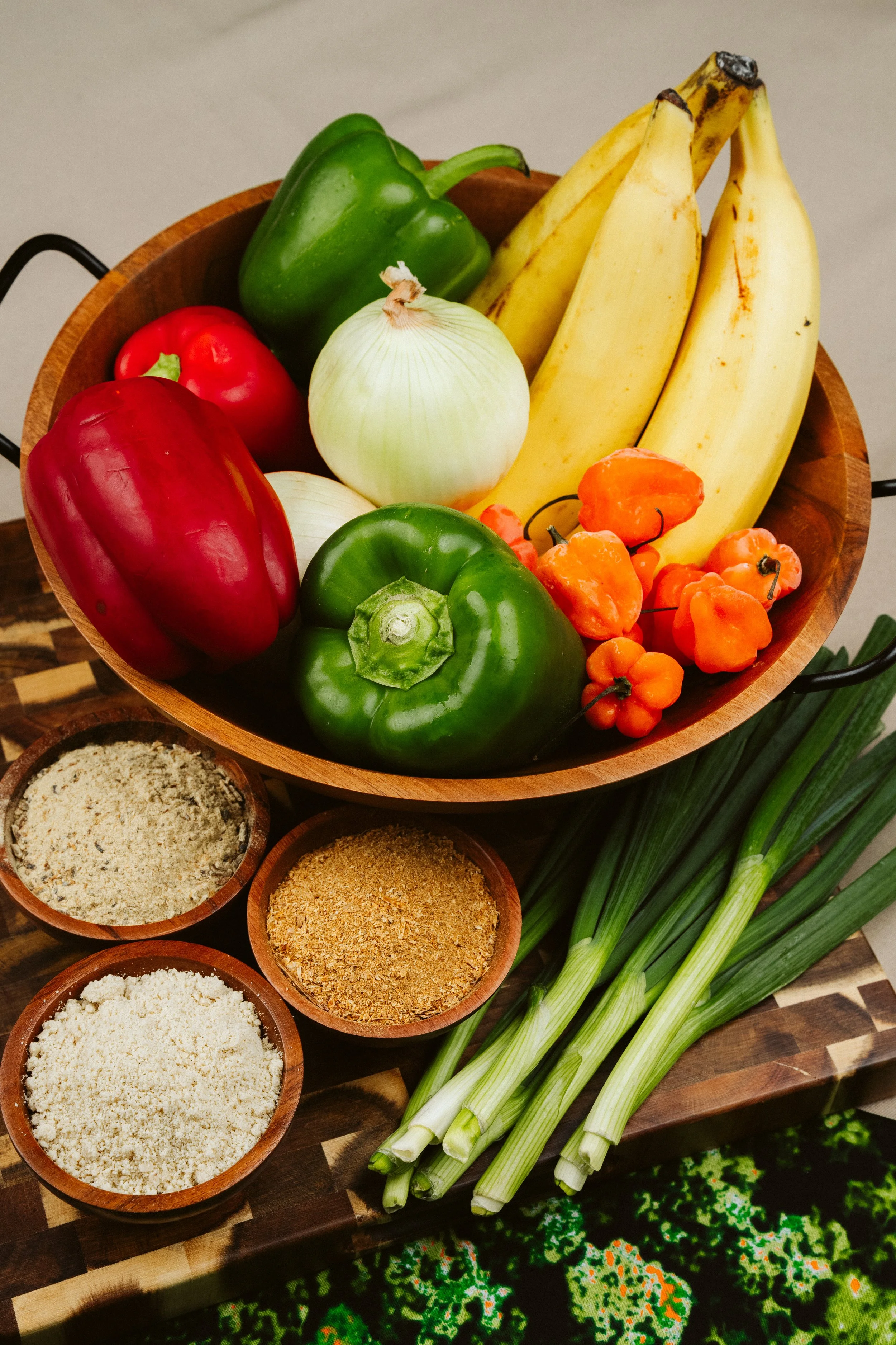 A wooden bowl filled with fresh green, red, and yellow bell peppers, a white onion, and bananas. Small bowls of various spices and herbs are nearby. Lemongrass stalks are laid out on the side.
