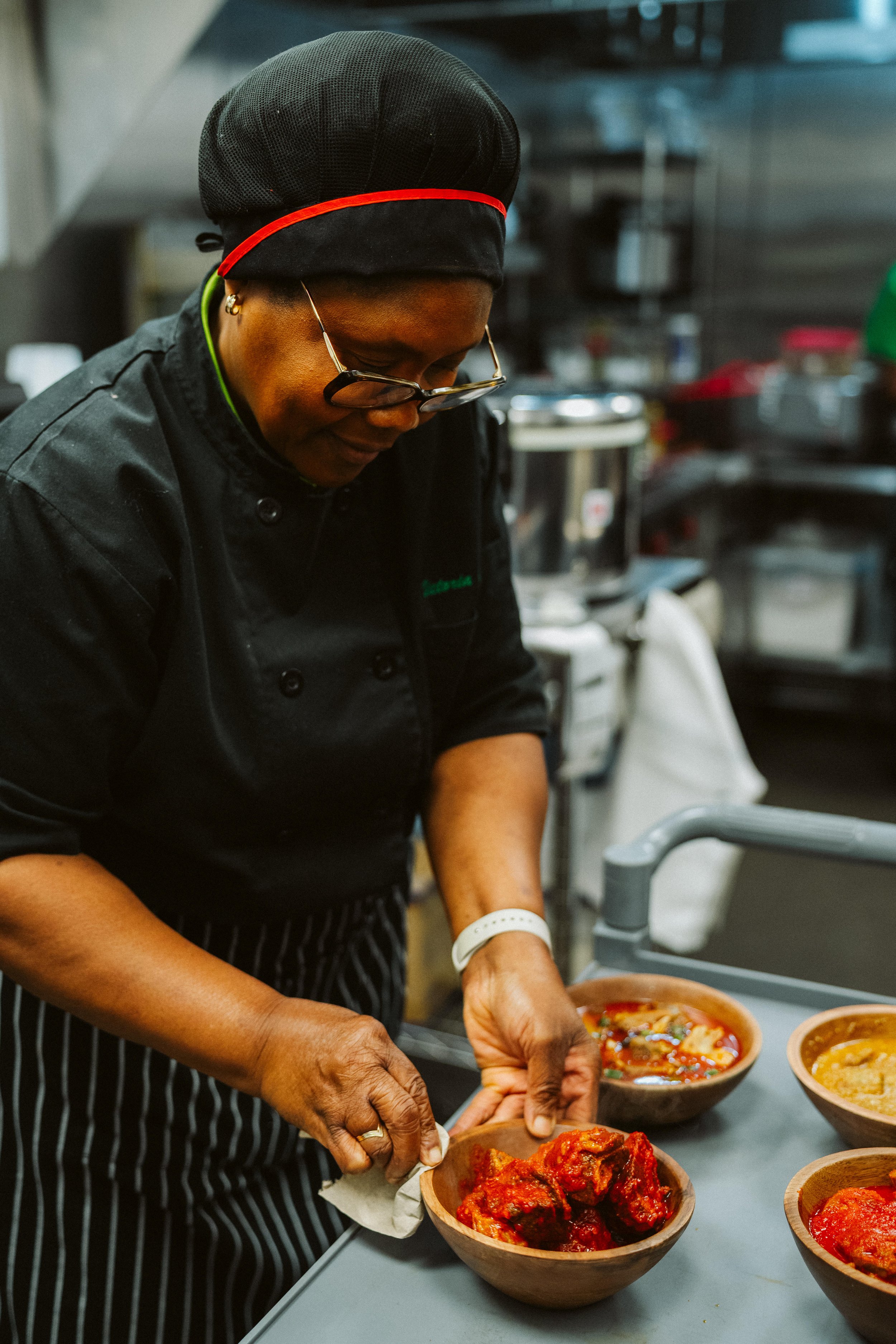 A woman wearing glasses, a black chef's coat, and a black and red hat, serving bowls of food in a kitchen.