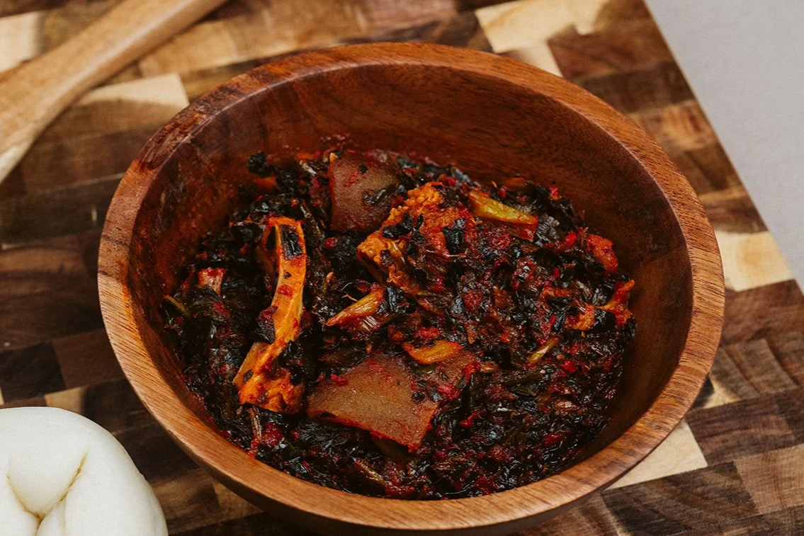 A wooden bowl containing a spicy, dark-colored vegetable stew with visible pieces of vegetables and tofu, placed on a wooden surface.
