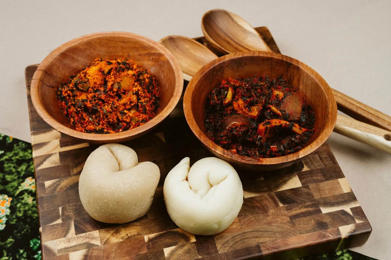 Two bowls of spicy, dark-colored, chunky food with vegetables on a wooden cutting board, alongside two steamed buns and wooden spoons.