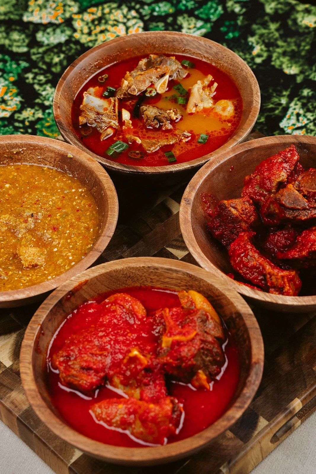 Five wooden bowls filled with different types of Indian curry and sauces, placed on a wooden tray with a patterned background.