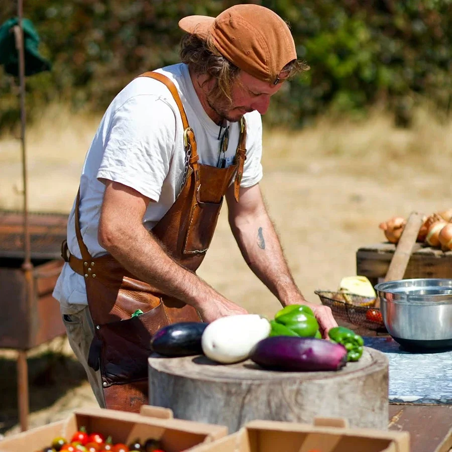 Man chopping vegetables outdoors, wearing a brown apron and a tan cap, with colorful eggplants and peppers on a wooden chopping block.