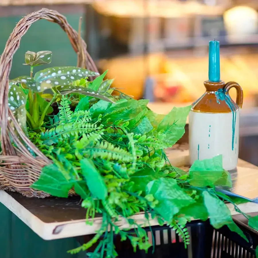 A basket filled with various green artificial plants sitting on a wooden table next to a brown and cream decorative bottle with blue paint dripping from its neck.