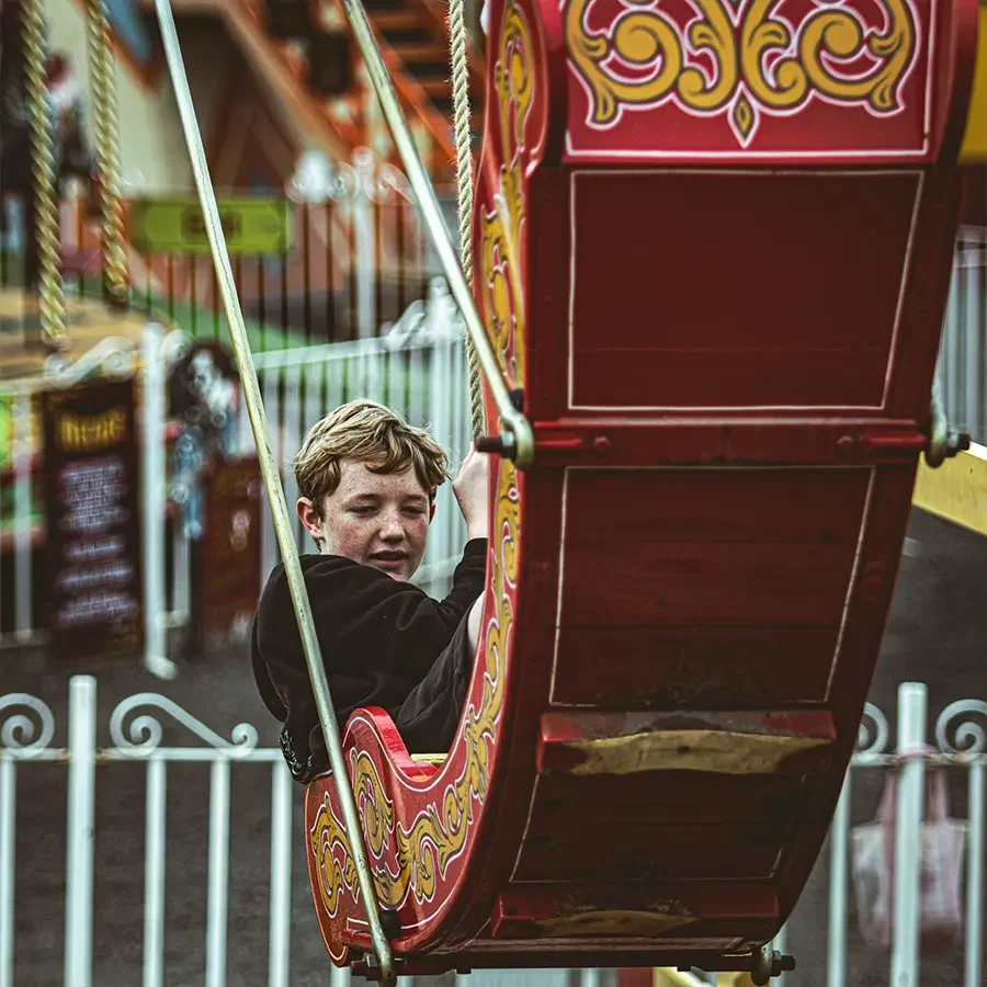 A young boy with blonde hair riding in a red and yellow ornate swinging boat at a funfair.