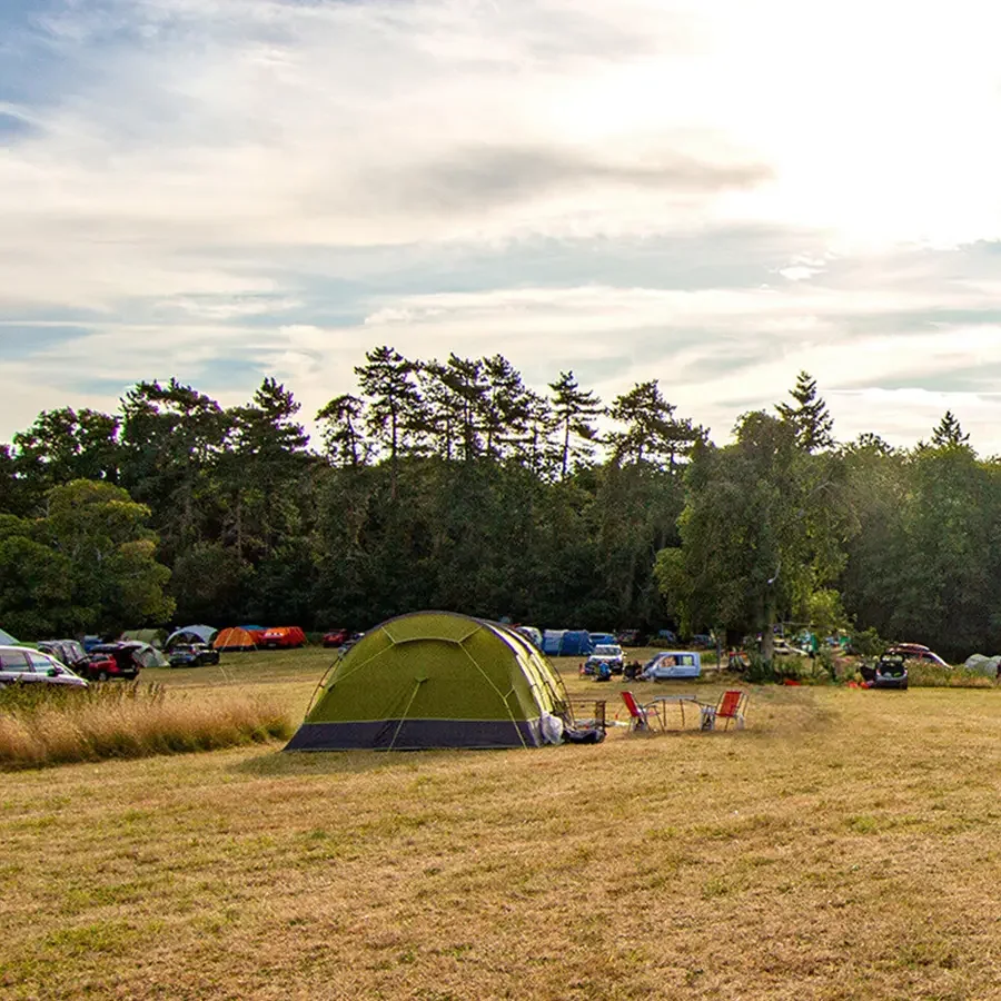 A camping scene with a large green and gray tent, two red chairs, and parked cars on a grassy field surrounded by trees under a partly cloudy sky.