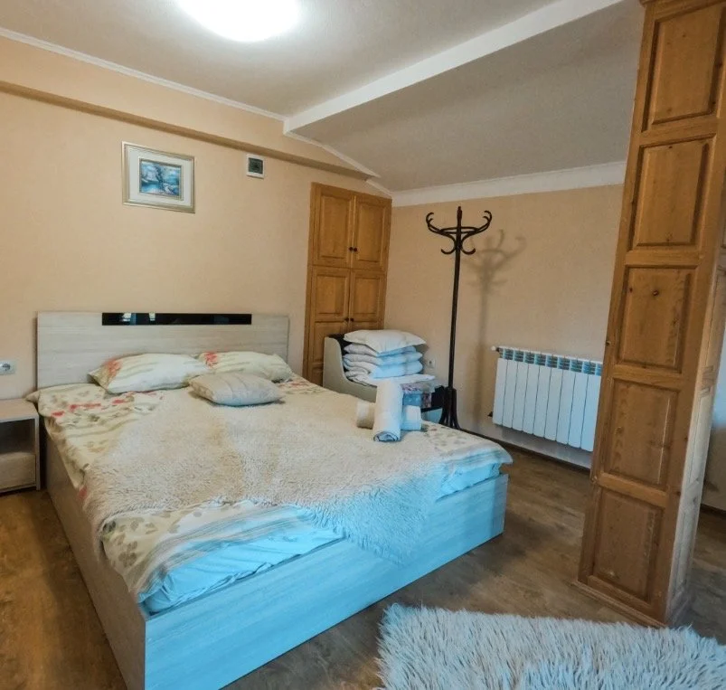 A bedroom with a wooden bed, stacked pillows, a rolled towel, a nightstand, a coat rack, wooden cabinets, a small framed picture on the wall, a radiator, and a fluffy rug on a wooden floor.
