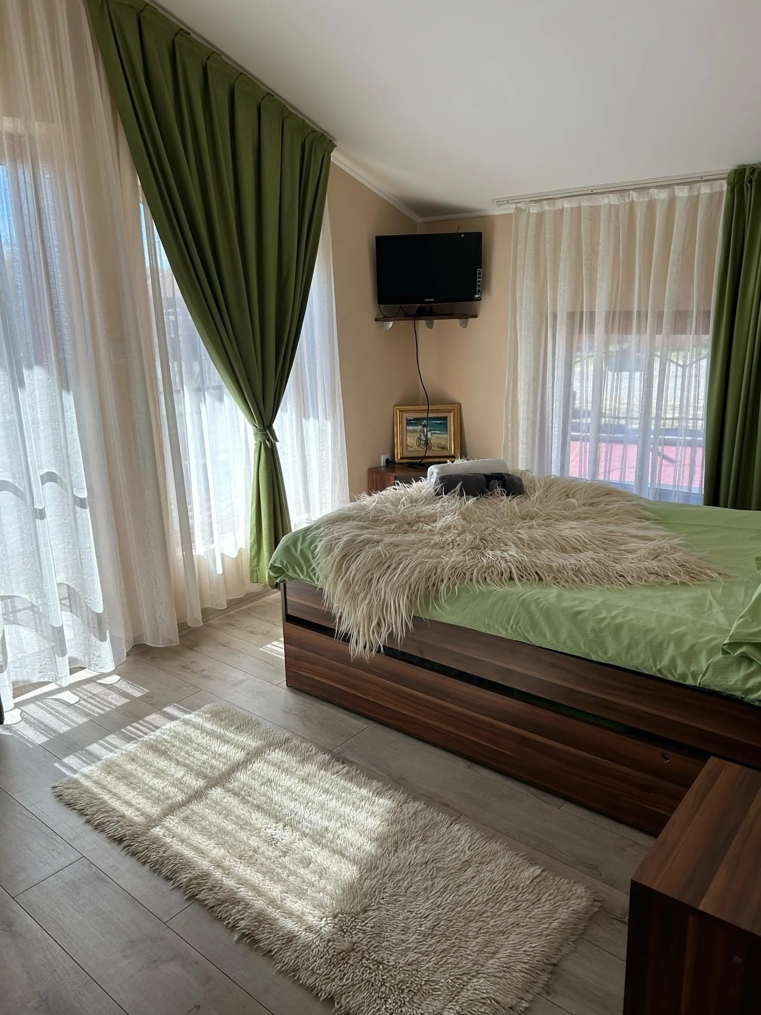 A cozy bedroom with a bed featuring a fluffy cream-colored blanket, green bedding, and a matching pillow. There are large windows with sheer white curtains and dark green drapes tied back. A small wooden nightstand with a framed painting and a TV mounted on the wall above it. The floor has light-colored wooden planks and a cream shaggy rug beside the bed.