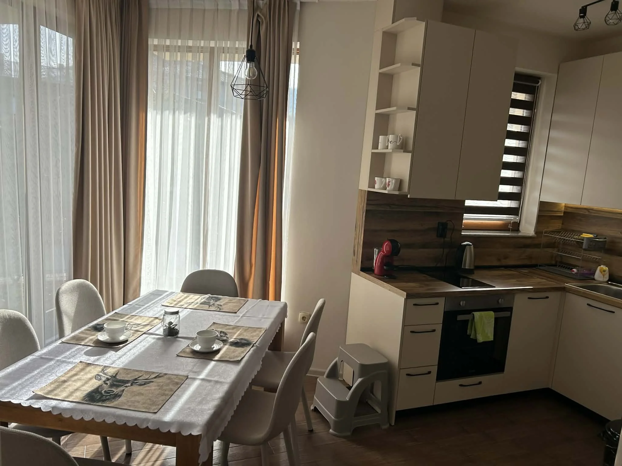 A dining area with a table set for four, a window with sheer curtains, and a modern kitchen with cabinets, a coffee machine, and a stove.