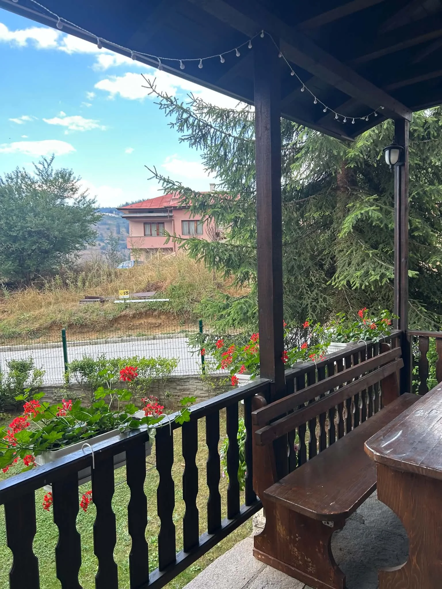 View from a covered porch with a wooden table and bench, red flowers in a planter, green trees, a house with a red roof, and a blue sky with clouds.