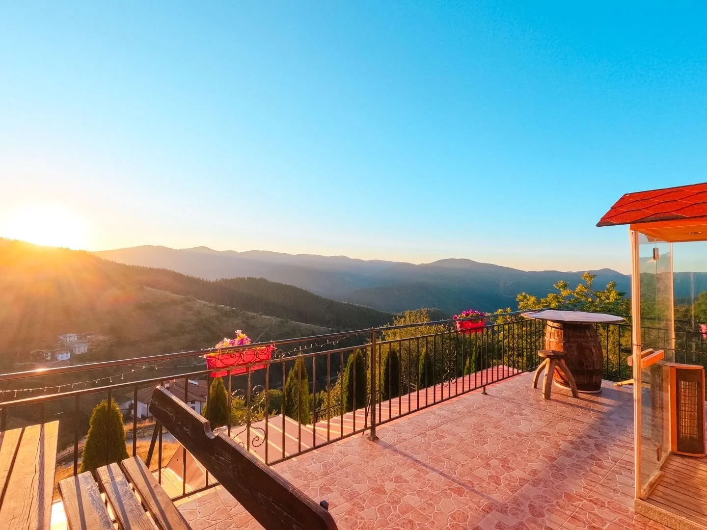 Sunset over mountain landscape viewed from a terrace with a black railing, pink flower boxes, wooden benches, and a small table with a barrel base, with a red-roofed corner and a glass enclosure.