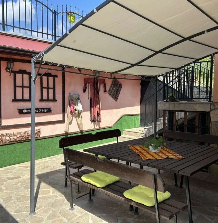 An outdoor patio area with a wooden table, benches with yellow cushions, and a large beige canopy overhead. The background shows a pink stucco wall with two small windows, a sign with Cyrillic script, hanging clothes, and stairs leading to an upper l