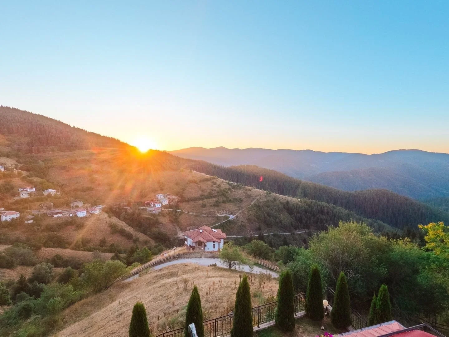 Sunset over rolling hills with scattered houses, trees, and a winding road in a mountainous landscape.