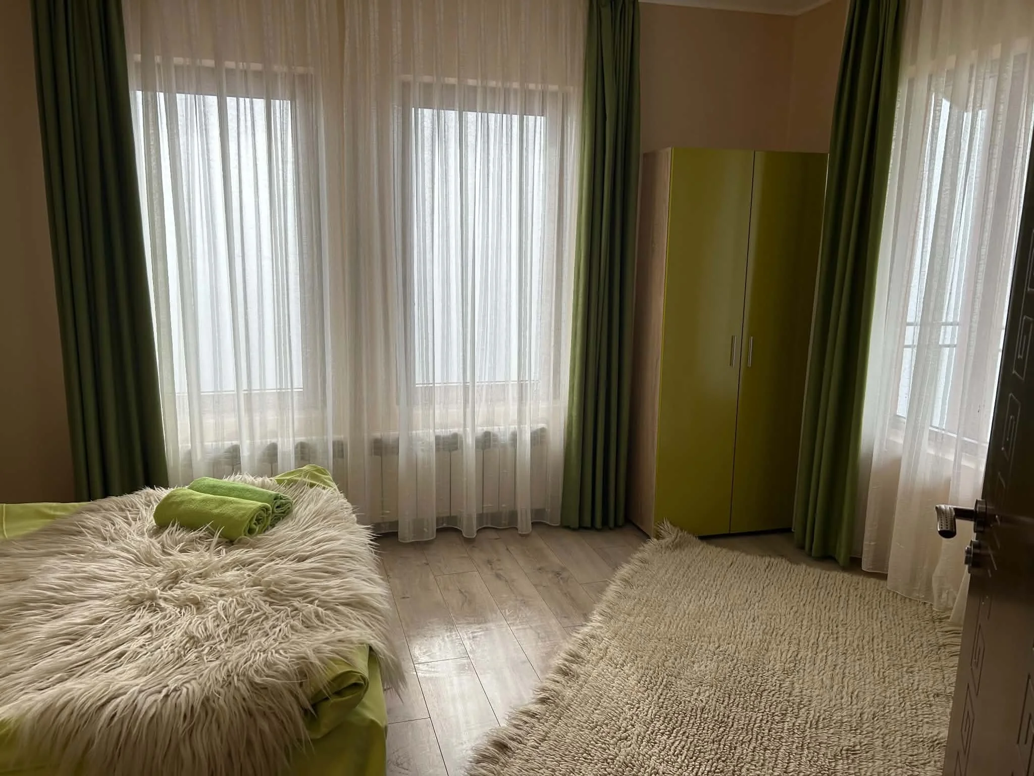 Room with large window with sheer white curtains and green drapes, yellow wardrobe, fluffy blanket with green pillows and towels, wooden floor, and beige rugs.