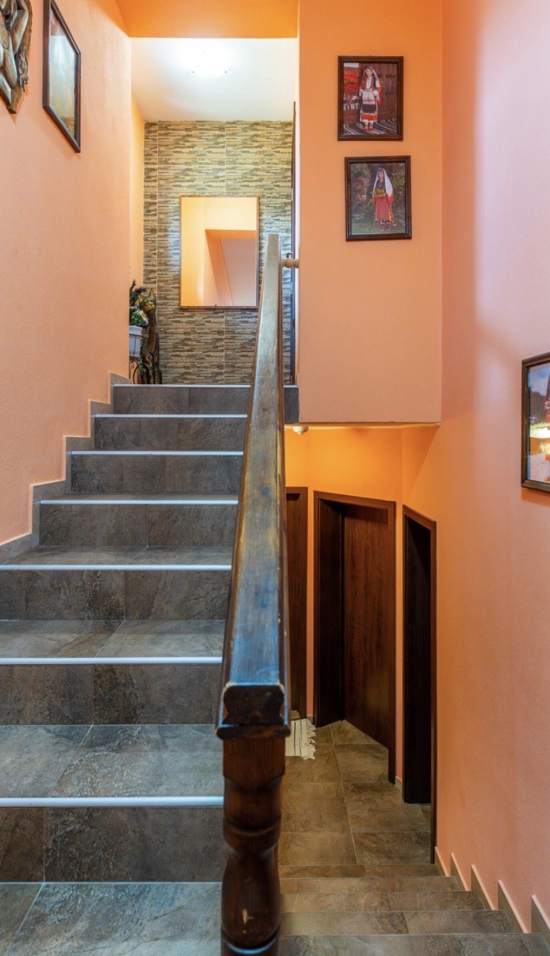 Inside a house, view of a staircase with tile steps and wooden handrail leading to the second floor. The walls are painted peach, and there are framed pictures hanging on the walls. At the top of the stairs, there's a textured wall with a rectangular