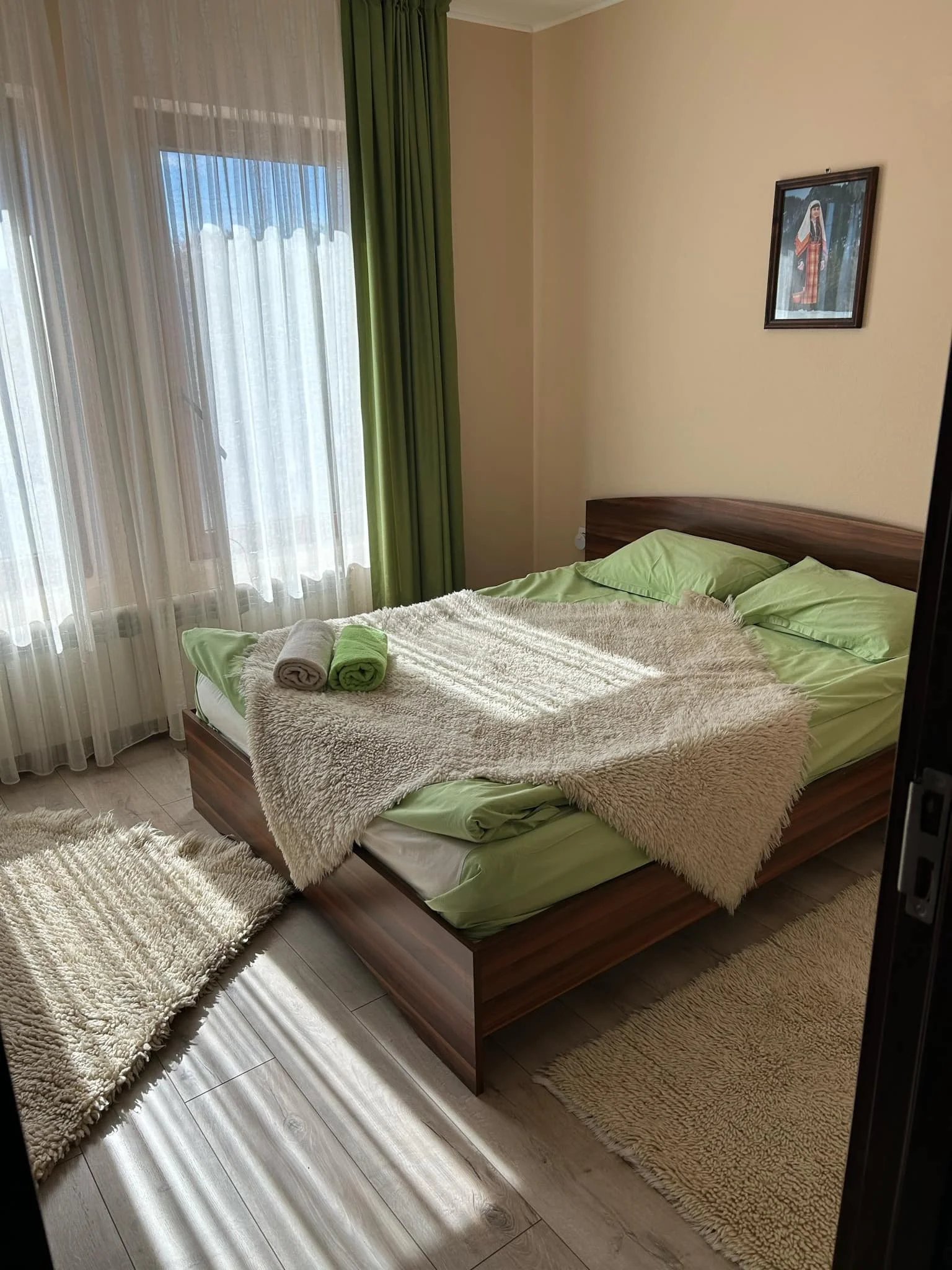 A bedroom with a bed covered in a beige fluffy blanket and green sheets, two rolled towels on top of the bed, a beige rug on the light-colored wooden floor, a window with sheer white curtains and green curtains, and a framed picture hanging on the wall.