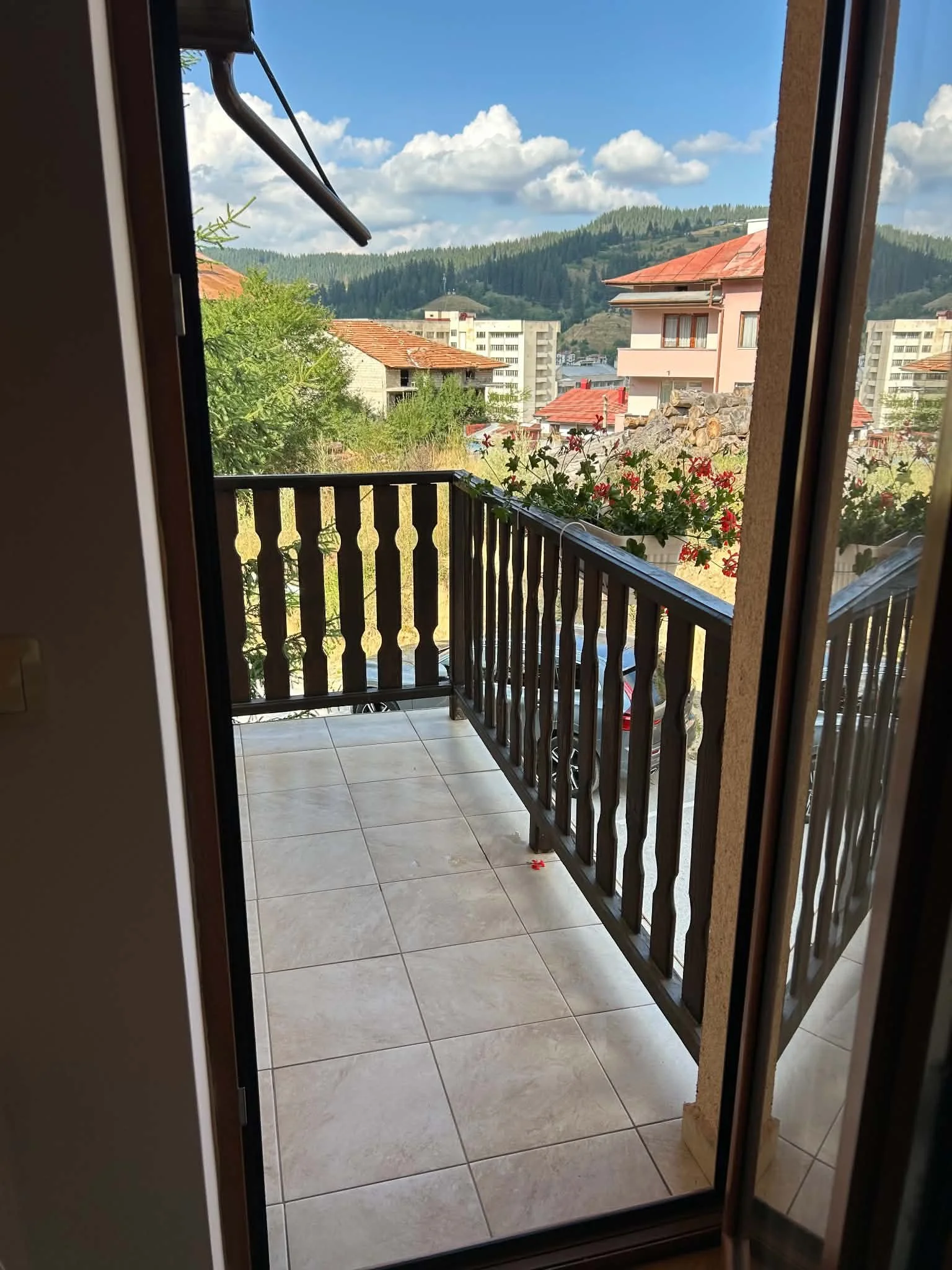 View from a balcony inside a room looking out through a sliding glass door to a small balcony and a scenic hillside. The scene includes a red-tilled roof house, mountain, trees, and blue sky with clouds.