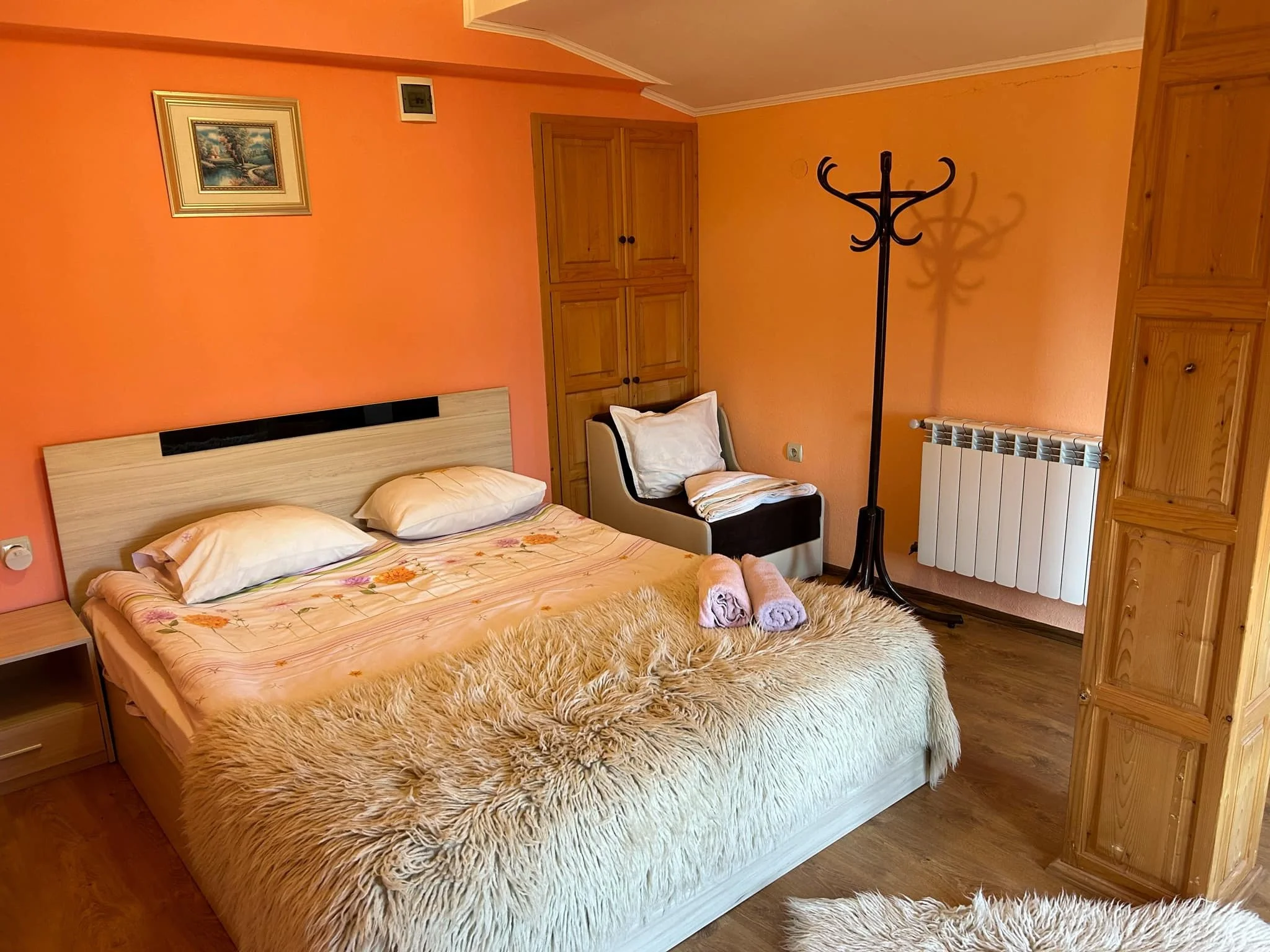 A cozy bedroom with orange walls, a bed with a floral bedspread, a furry beige rug, wooden cabinets, a black coat rack, a chair with pillows and folded blankets, a picture on the wall, and a radiator.
