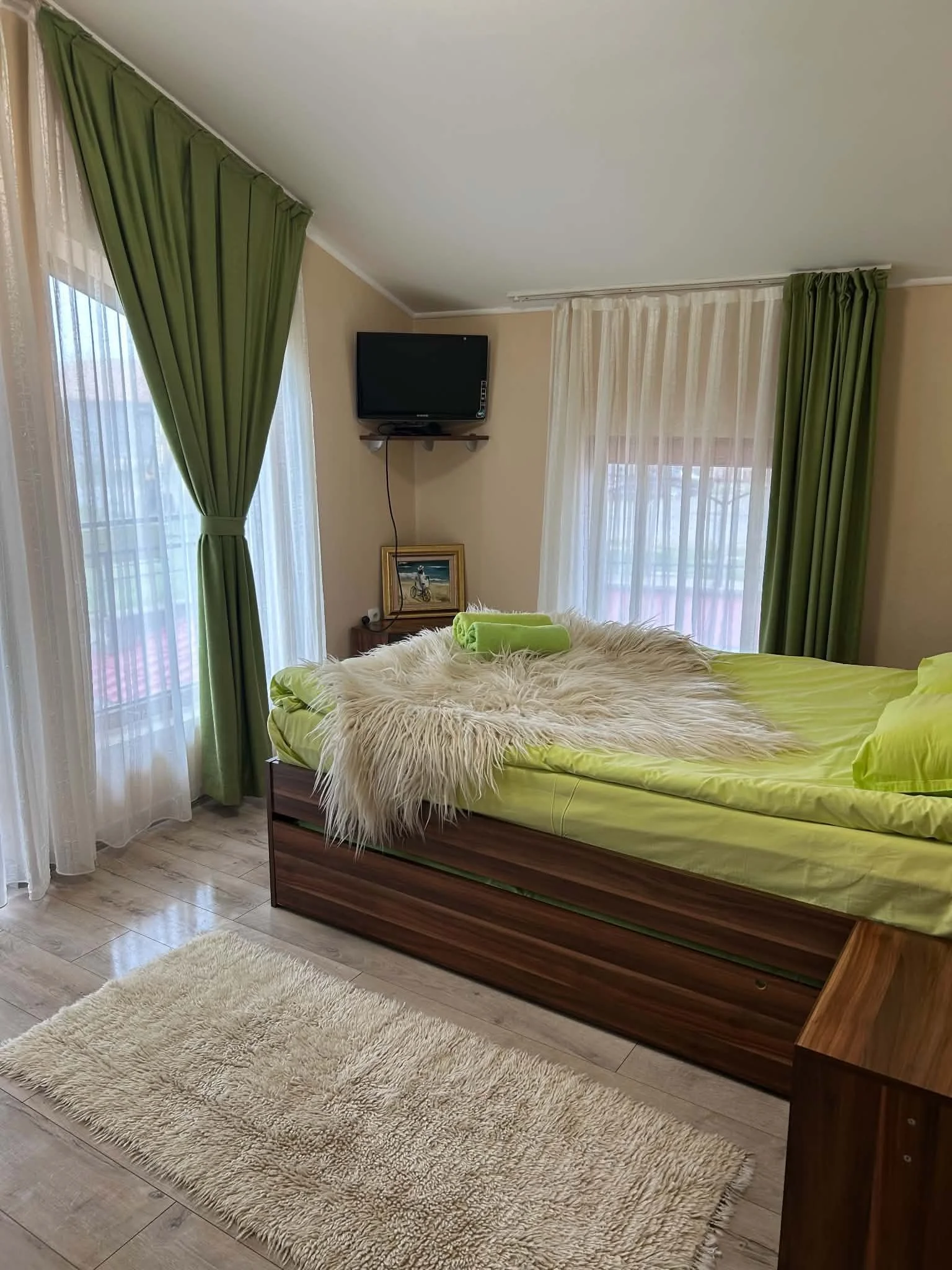 A bedroom with a bed covered in green sheets and a fluffy white rug on the floor. Two large windows with sheer curtains and green drapes, and a small flat-screen TV mounted on the wall.