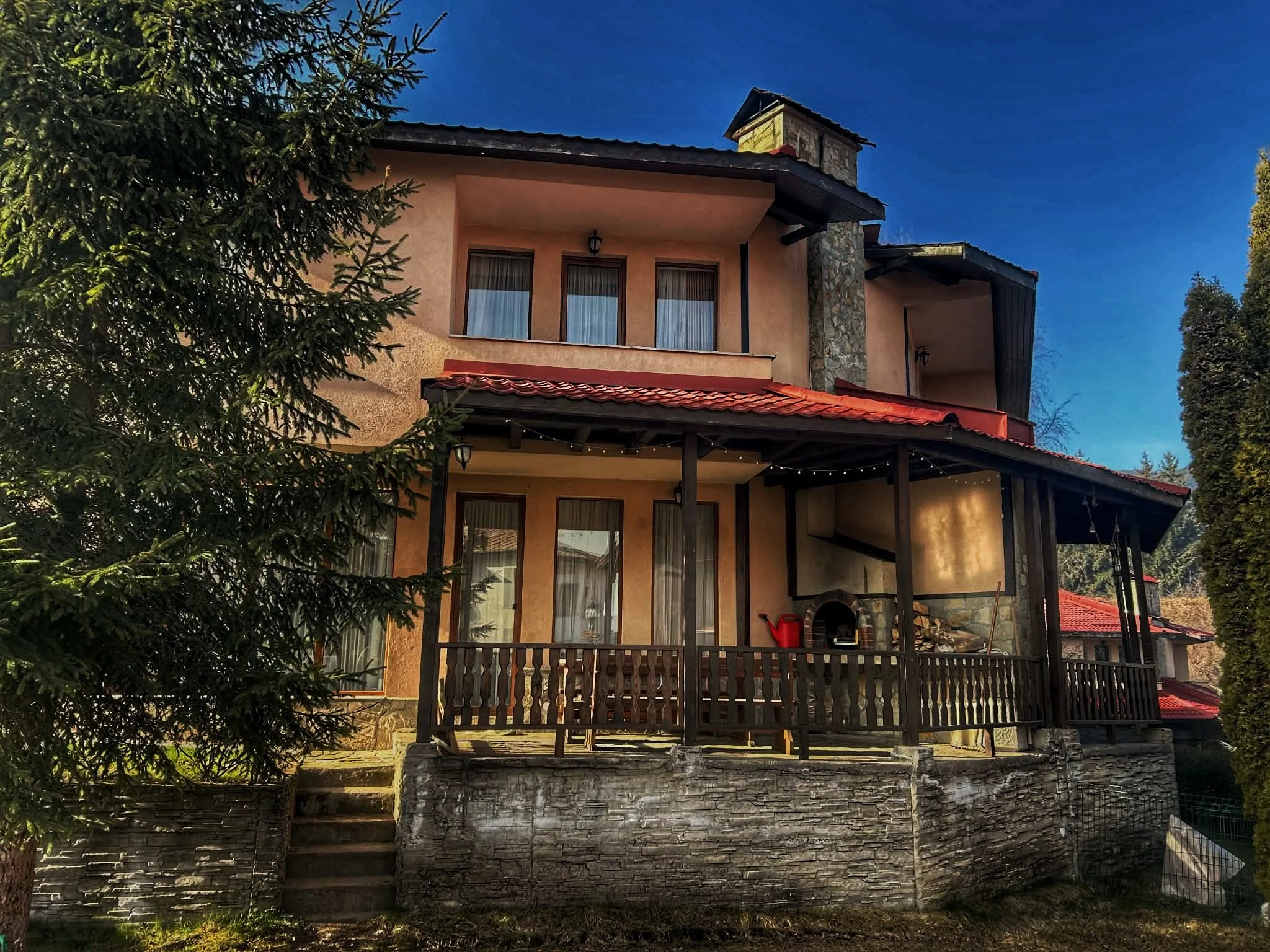 Two stories house with beige stucco walls, dark roof, stone chimney, large windows with curtains, and a wooden deck with string lights, surrounded by trees.