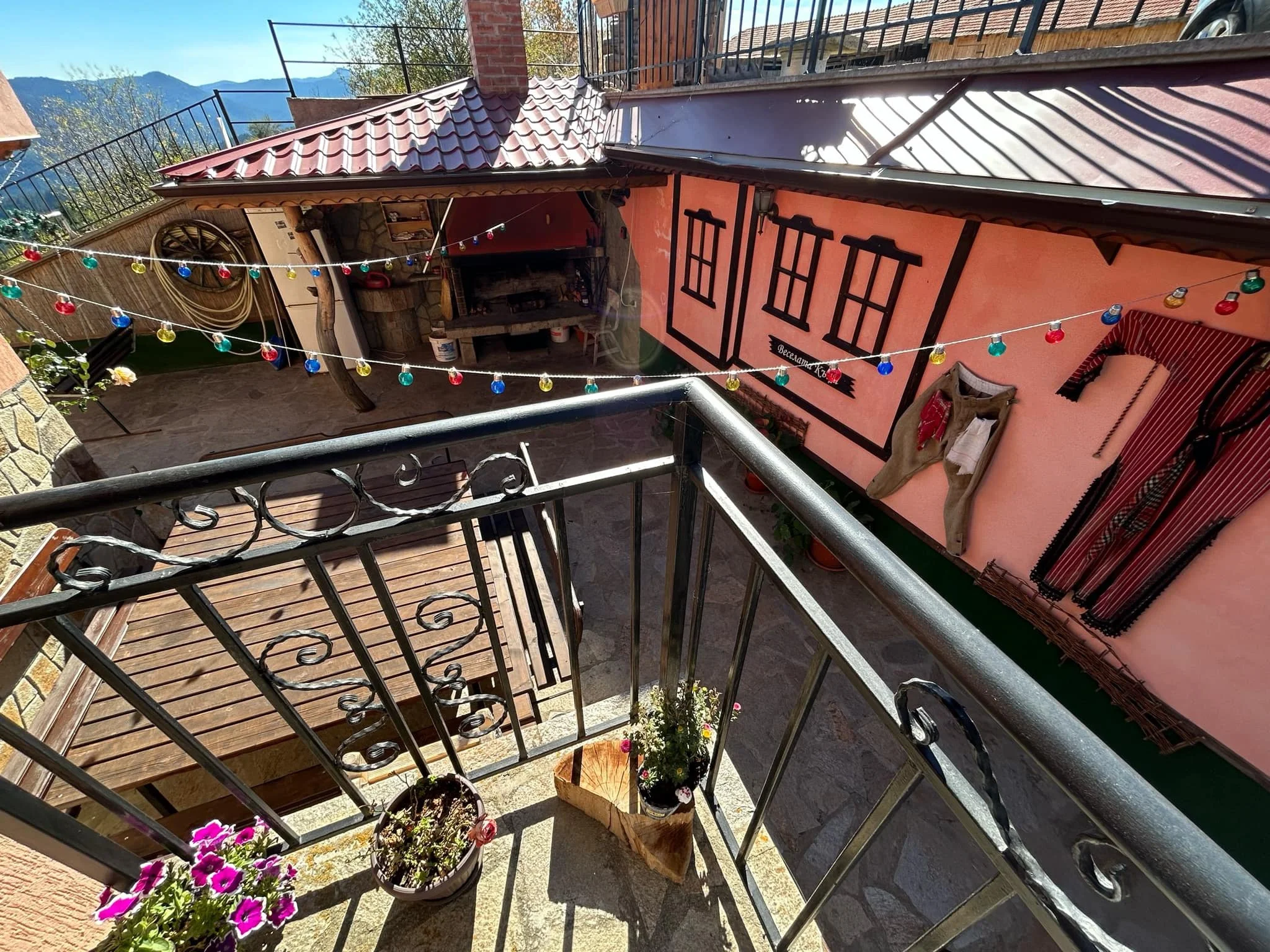 A view from a balcony looking down at a small outdoor patio with potted flowers, a wooden table with benches, and colorful string lights, with mountains visible in the background.