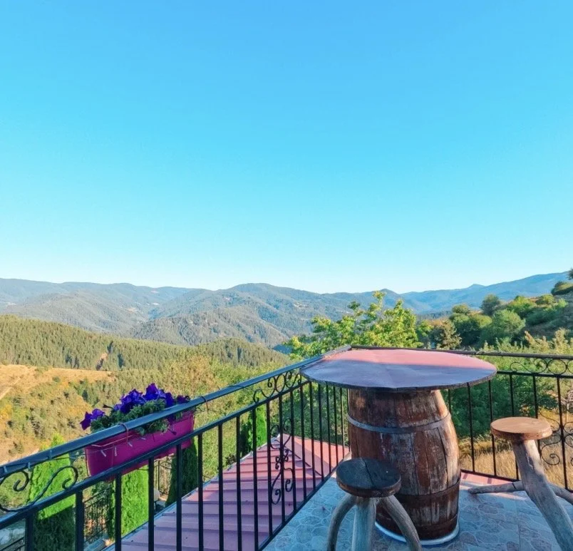 A balcony overlooking mountains with a barrel-shaped table, two small stools, and a flowerpot with purple flowers.
