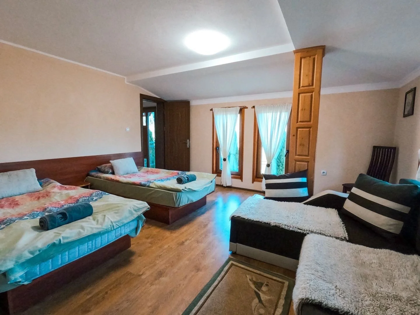 A bedroom with two beds, a sofa, and a wooden interior design. The beds have floral patterned bedspreads and rolled towels. There are two windows with sheer white curtains, a wooden door, and wooden paneling. The floor is hardwood, and there is a small rug with a dog illustration.