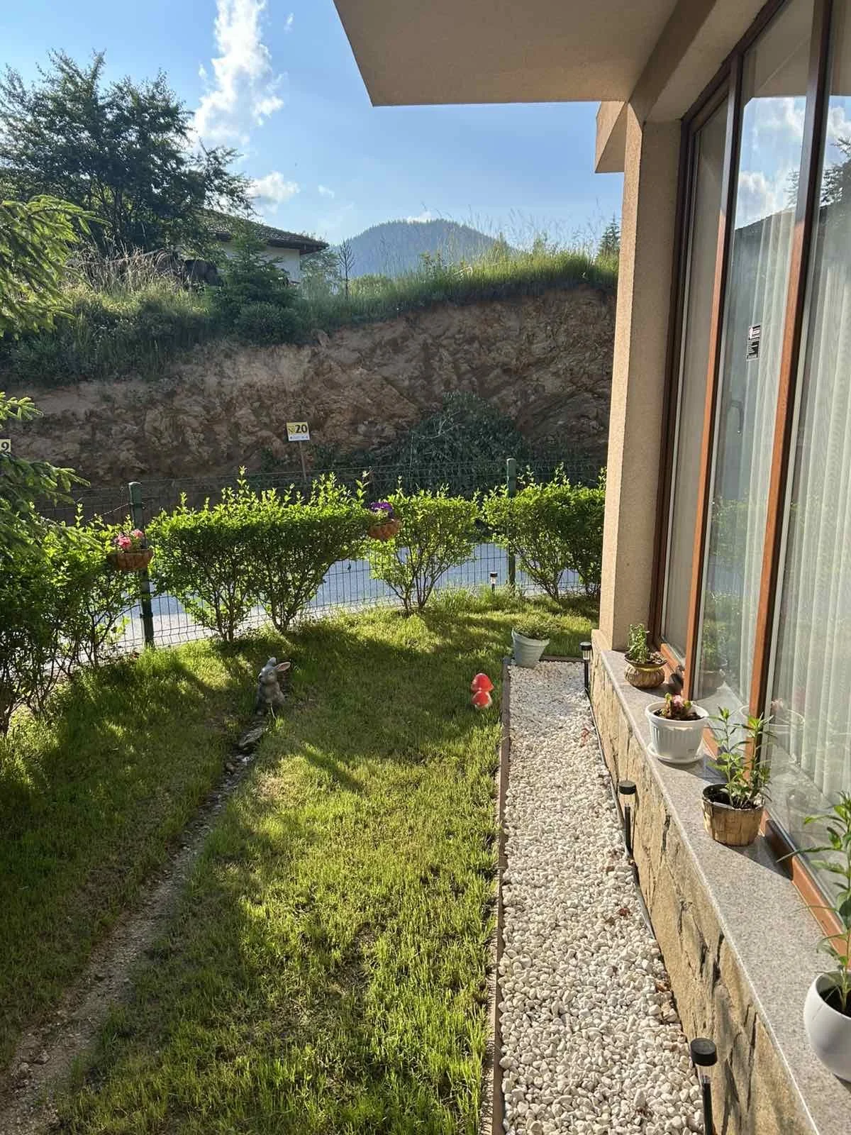 A small garden patio with decorative plants on the right, a lush green lawn on the left, and a hedge and mountains in the background under a bright blue sky.