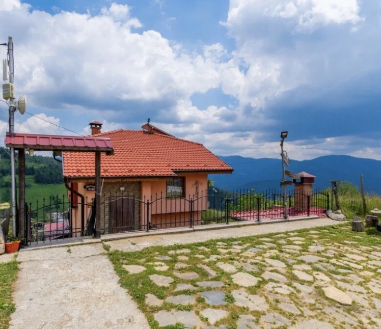 A small house with a red tiled roof and peach-colored walls, surrounded by a black metal fence, on a grassy hill with cobblestone pathway, mountains in the background, and a partly cloudy sky.