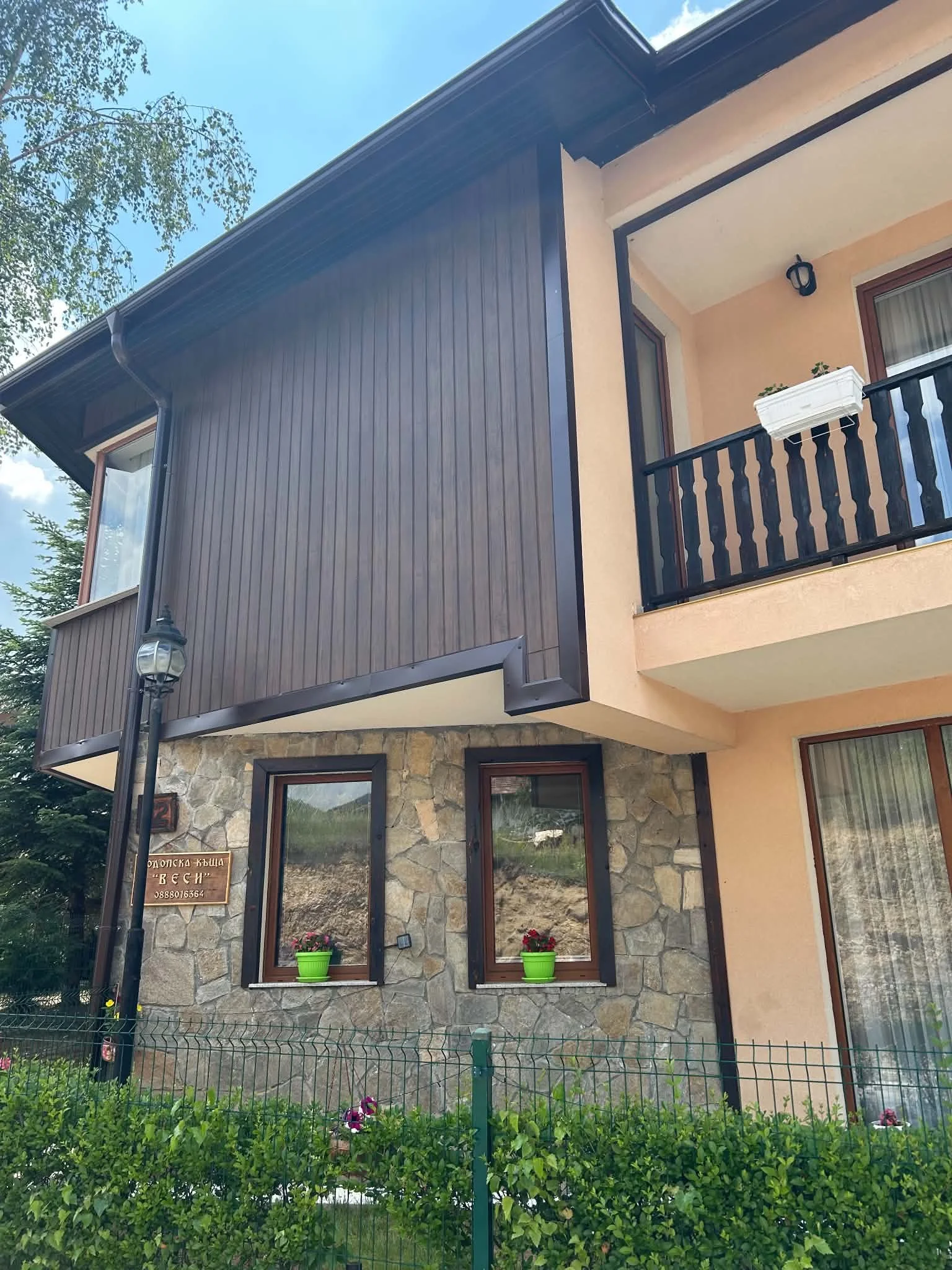 A two-story house with stone and peach-colored exterior walls, featuring a small balcony with black railing, green potted plants, windows with brown frames, and a sign in Cyrillic script on the stone wall, surrounded by greenery and a green fence.