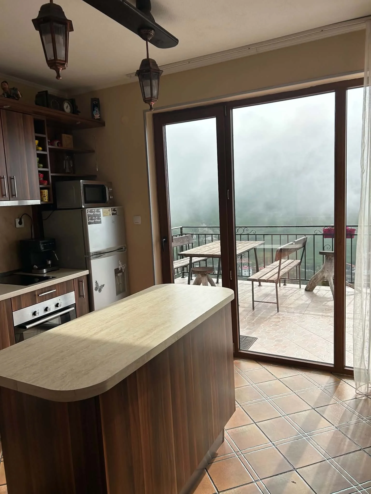 The image shows a kitchen with a view of a balcony outside through a glass door. Inside, there is a wooden kitchen counter, a microwave, a refrigerator, and cabinets. The balcony has outdoor furniture, including a table, chairs, and a bench, with a r