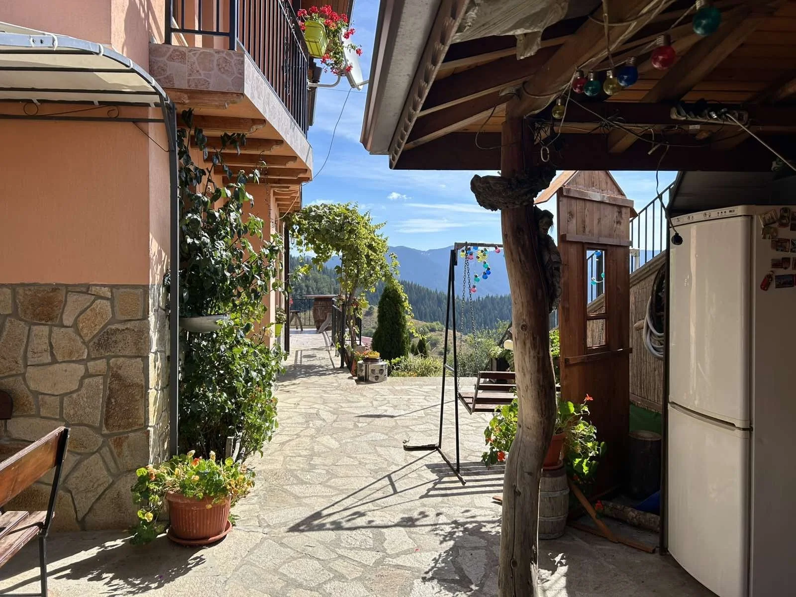 A stone-paved outdoor patio area with potted plants and outdoor furniture, overlooking distant mountains and a blue sky, with a red-flowered balcony above.