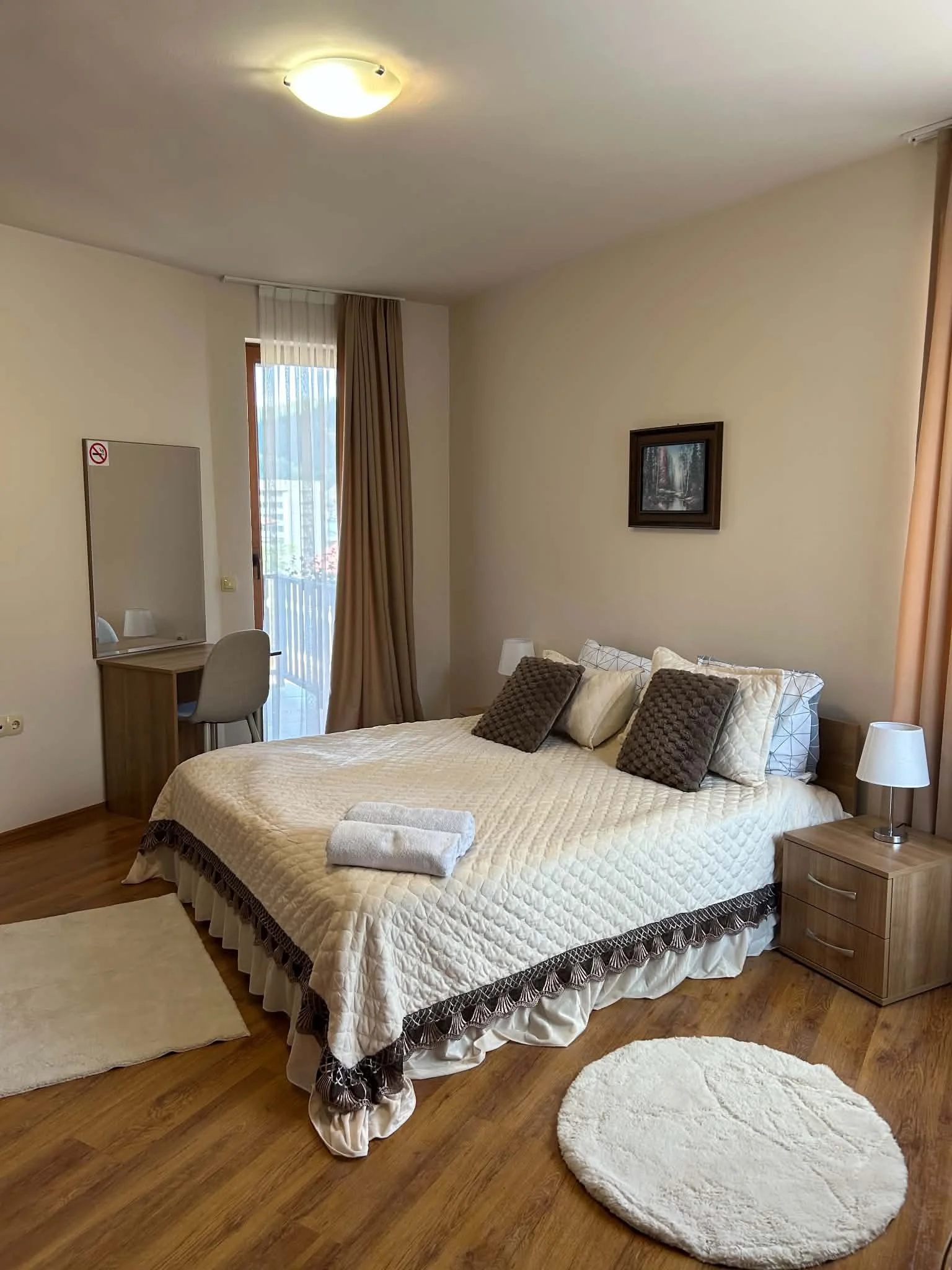 A bedroom with a bed covered in a light quilt, topped with four brown and light-colored pillows, and a folded towel on top. To the right of the bed is a wooden nightstand with a small lamp. The room has hardwood floors, a beige carpet, and a round be