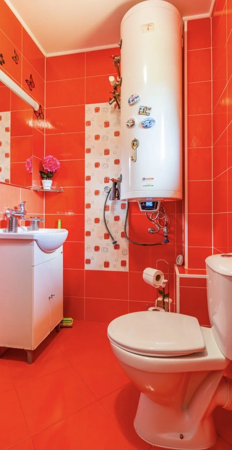 A small bathroom with red tiled walls and floor. There is a white toilet, a white sink with a cabinet below, and a white water heater mounted on the wall. The bathroom has pink artificial flowers on a small glass shelf and some decorative magnets on 