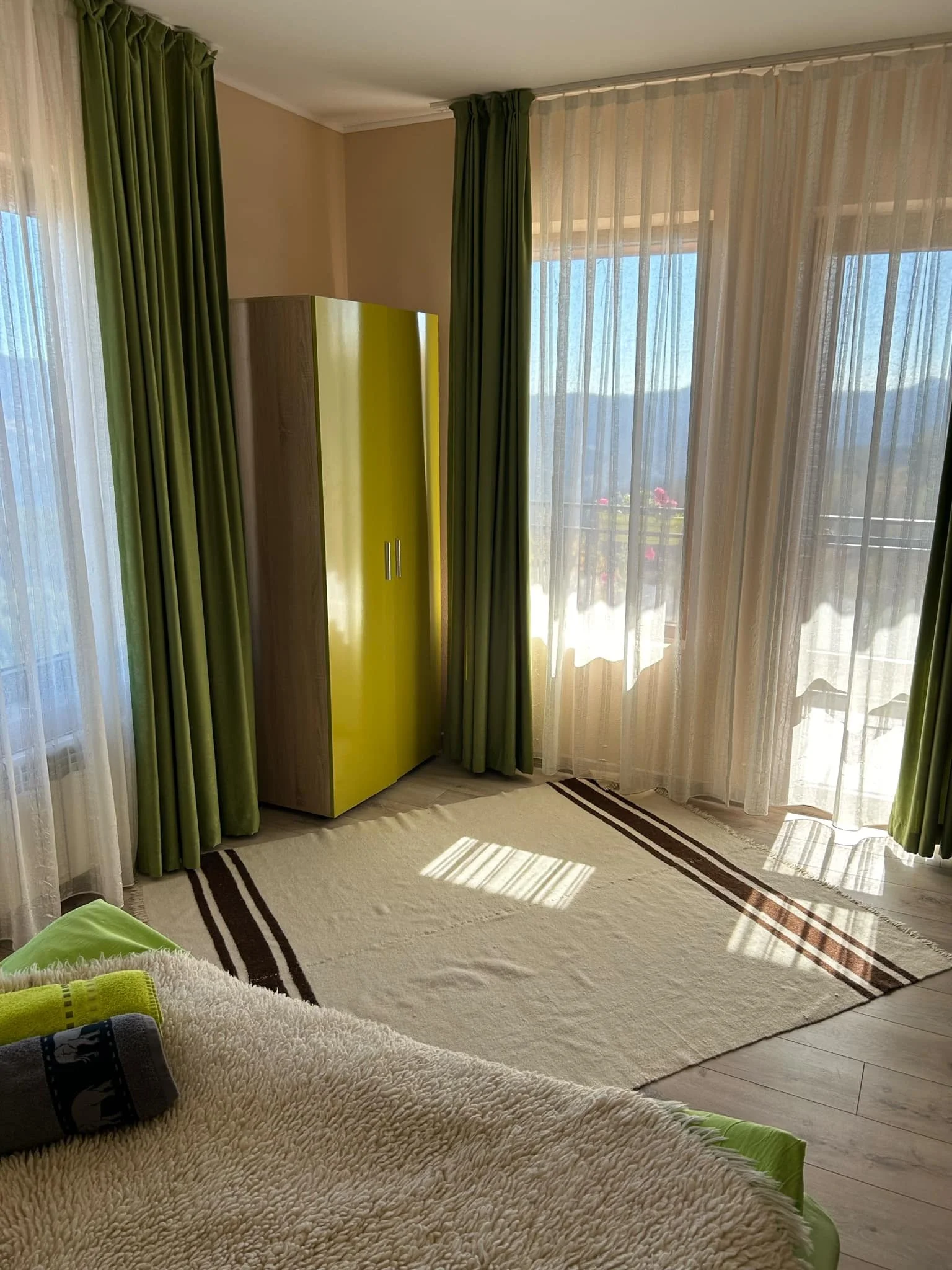 Bright room with large windows covered by sheer curtains and green drapes, beige walls, and light-colored wood flooring. There is a bed with a shaggy beige blanket and a green pillow in the foreground, and a yellow wardrobe near the window. Sunlight 