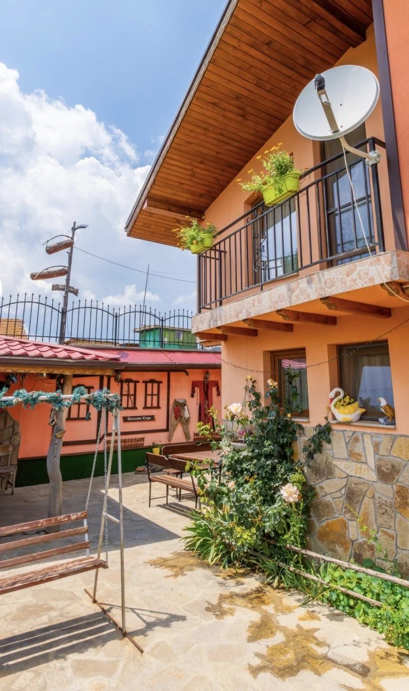 A cozy patio with wooden swing, garden plants, and decorative swan figure in front of a peach-colored house with a balcony and satellite dish, under a partly cloudy sky.