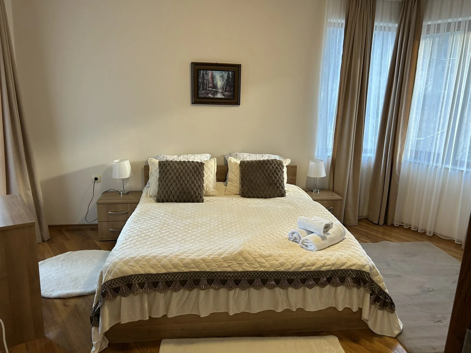 A cozy bedroom with a large bed, four pillows, two lamps on bedside tables, a framed landscape picture on the wall, and curtains covering windows, with towels placed on the foot of the bed.