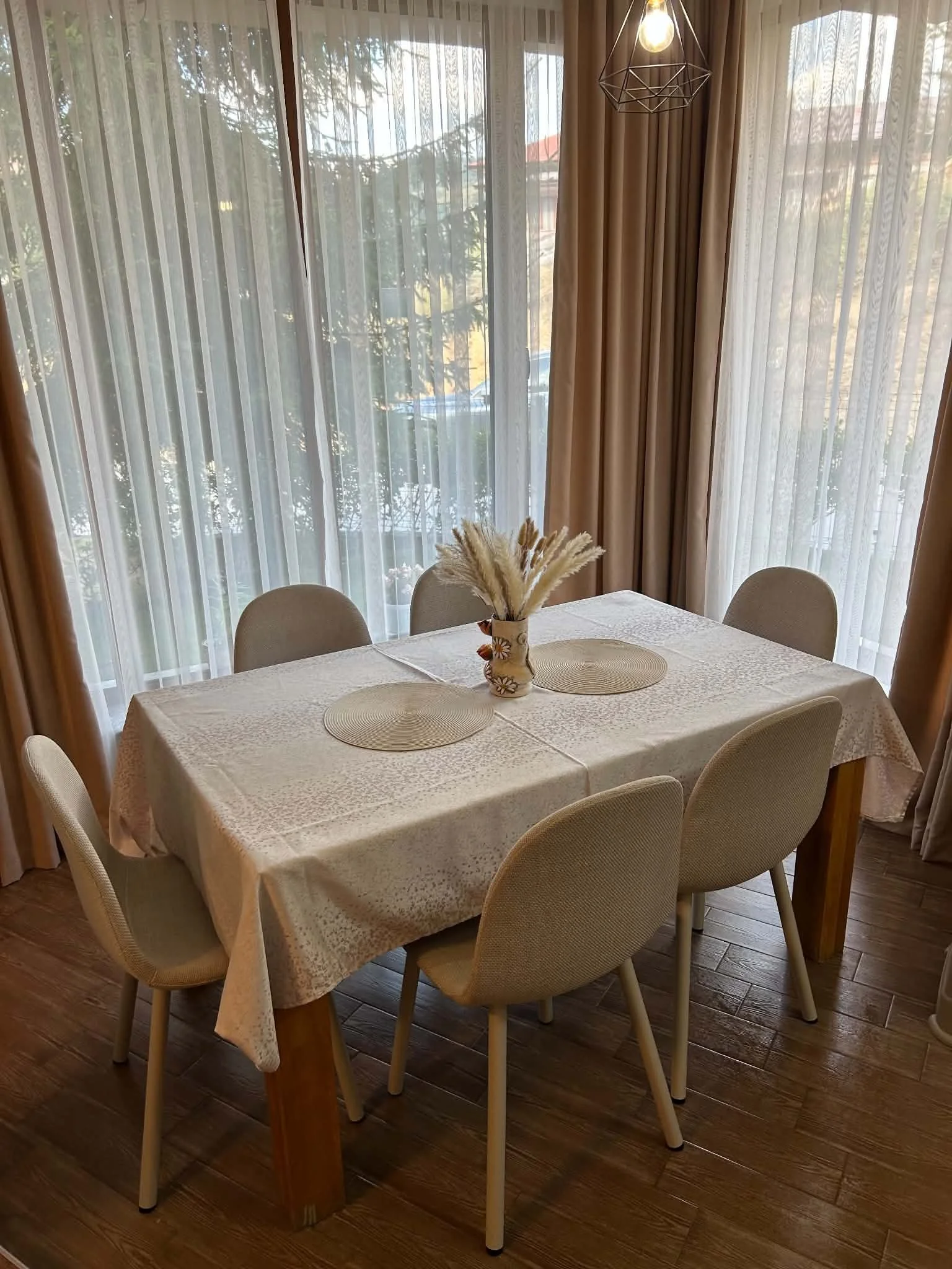 Dining table with a white tablecloth and six beige chairs, a vase with dried flowers, and a background of large windows with sheer curtains and brown drapes, allowing natural light to fill the room.