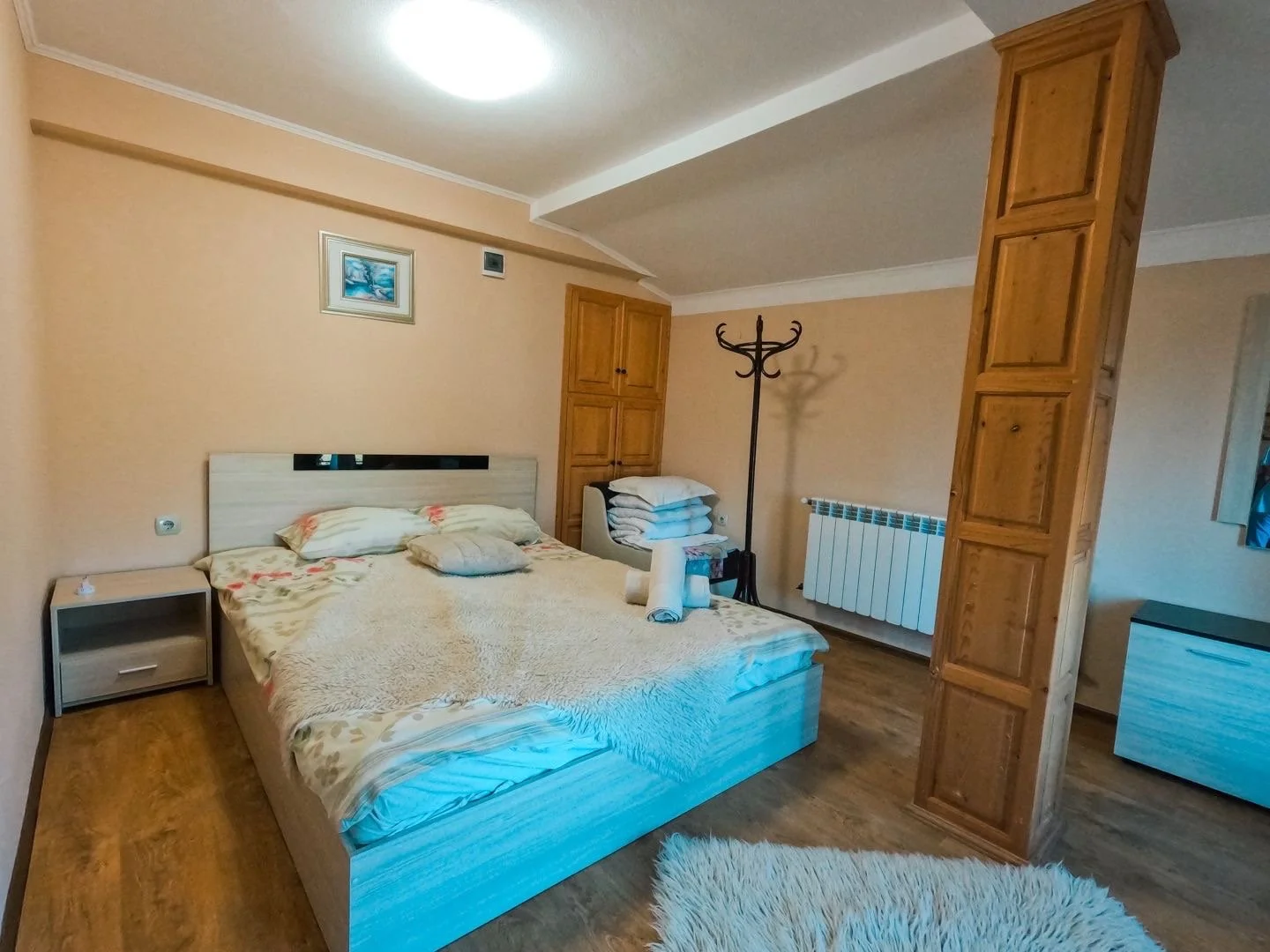 A bedroom with a double bed, a nightstand, and a stack of folded blankets. The room has wooden cabinets, a coat rack, a radiator, and a picture on the wall. A fluffy rug is on the wooden floor.