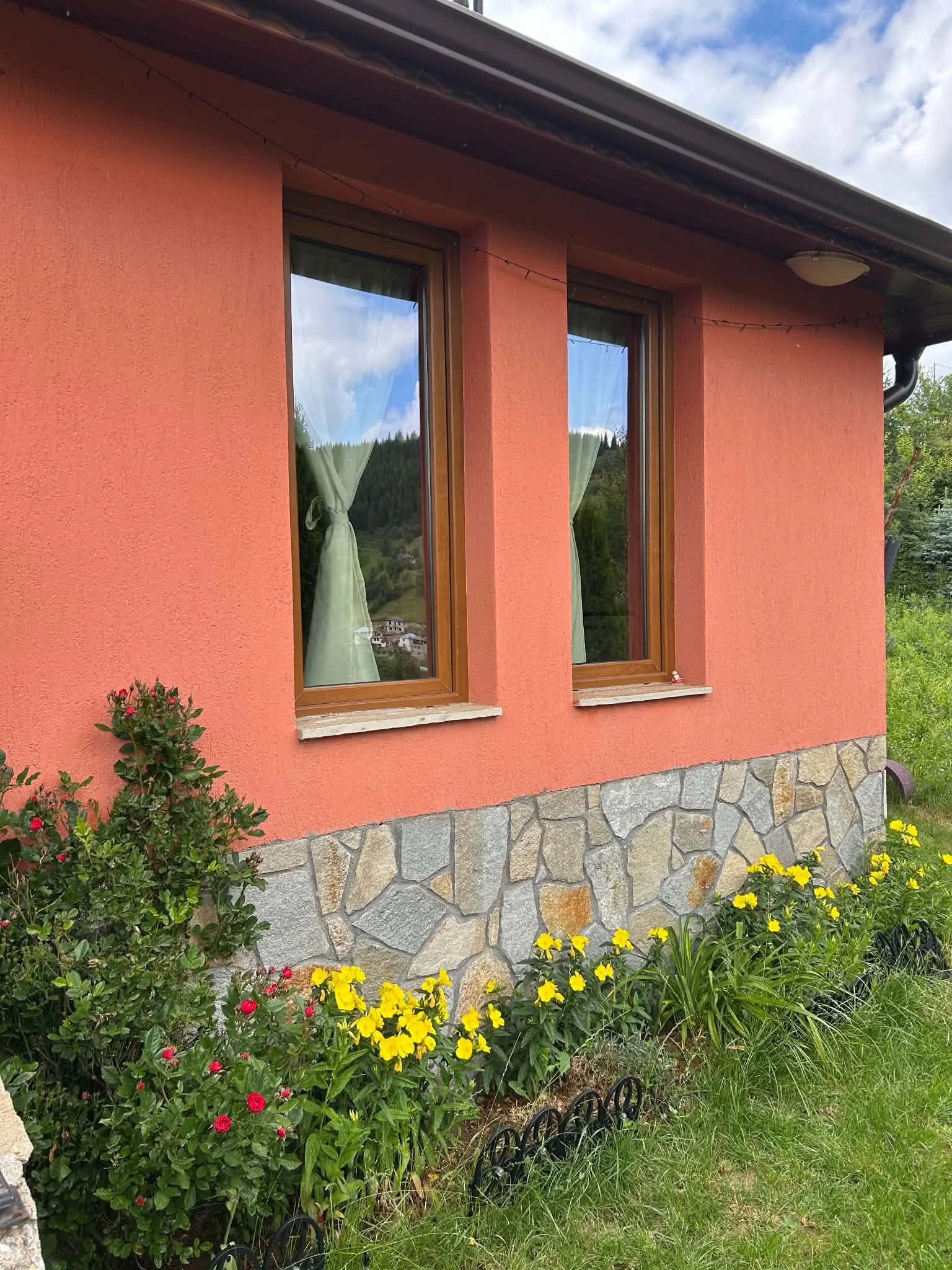 A close-up view of the exterior of a house with coral-textured walls, stone foundation, large rectangular windows with wooden frames, curtains visible inside, and a lush garden with colorful flowers and green plants.
