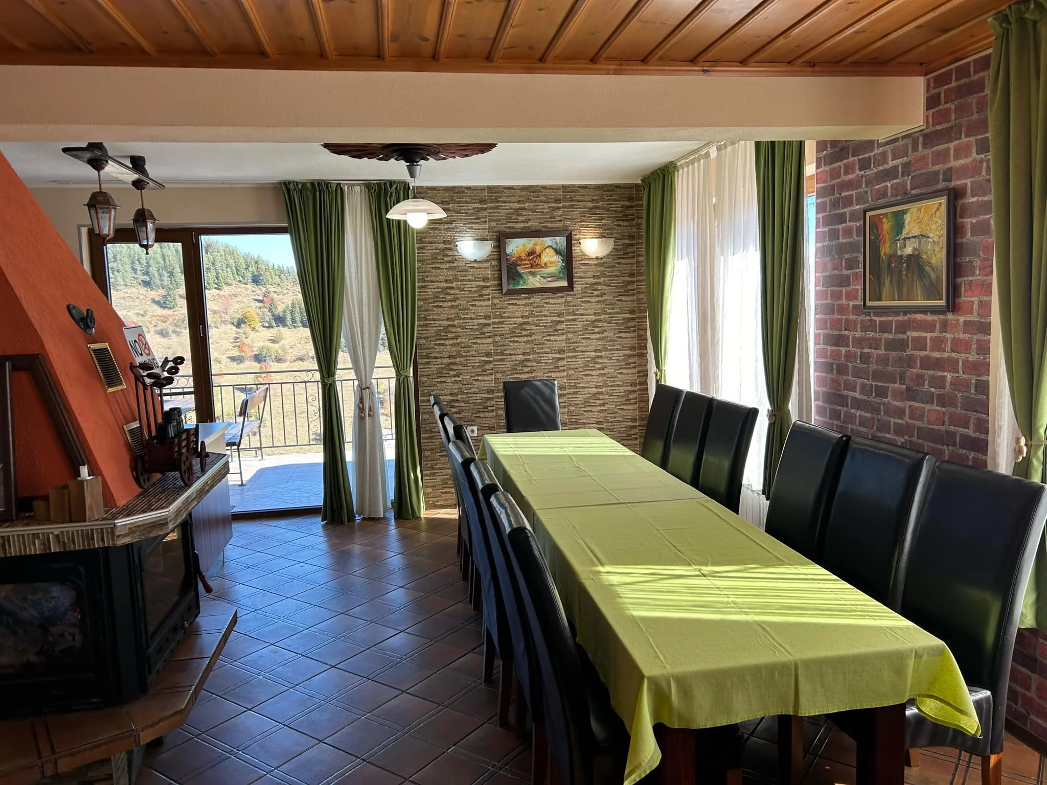 Interior of a dining area with a large window showing a scenic view of trees and a hillside. The room features green curtains, a brick accent wall, and framed paintings. There is a long dining table with a green tablecloth and black chairs, alongside