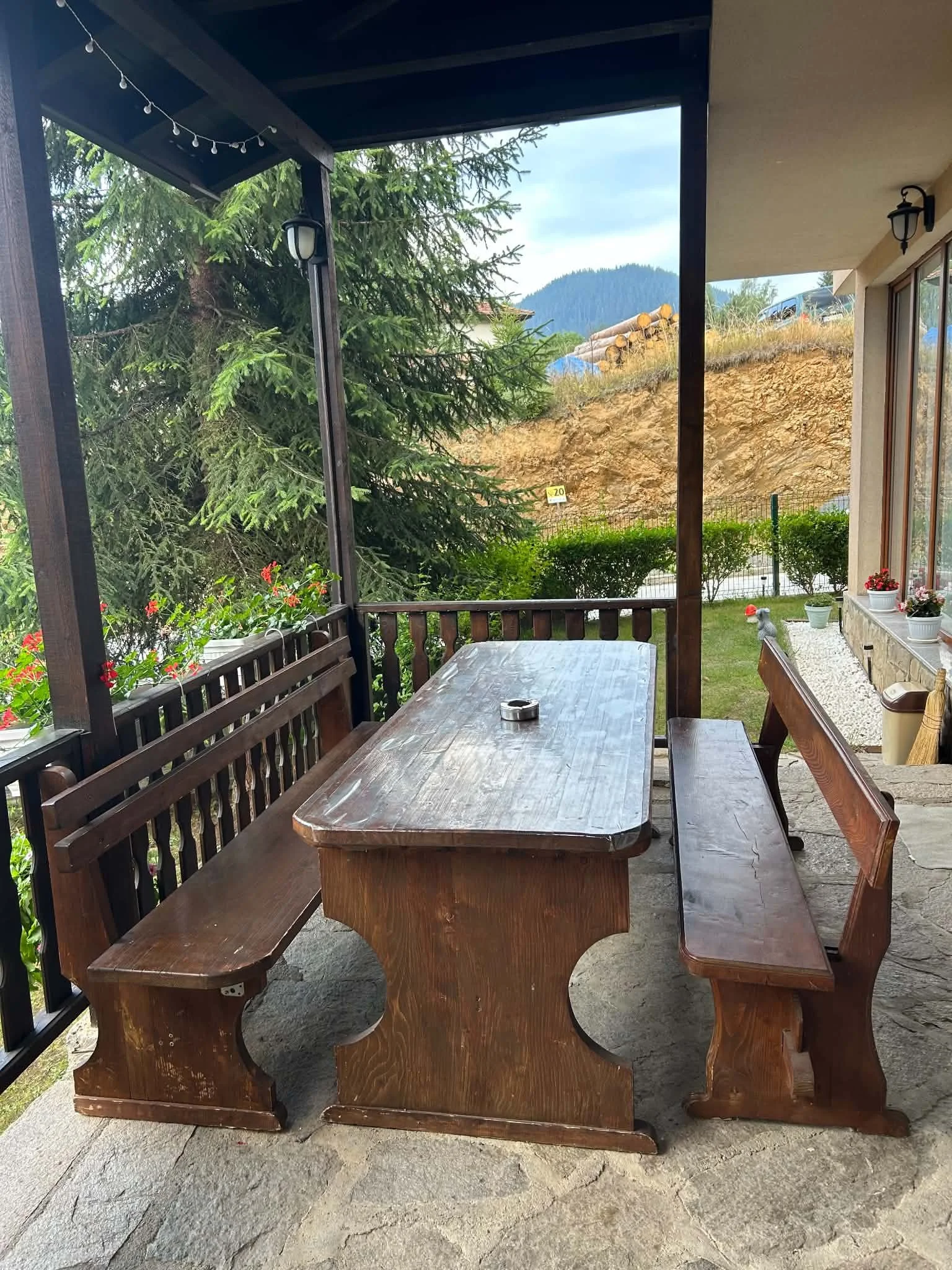 A wooden outdoor table and benches on a stone patio, with a view of trees, a hillside, and a distant mountain in the background.