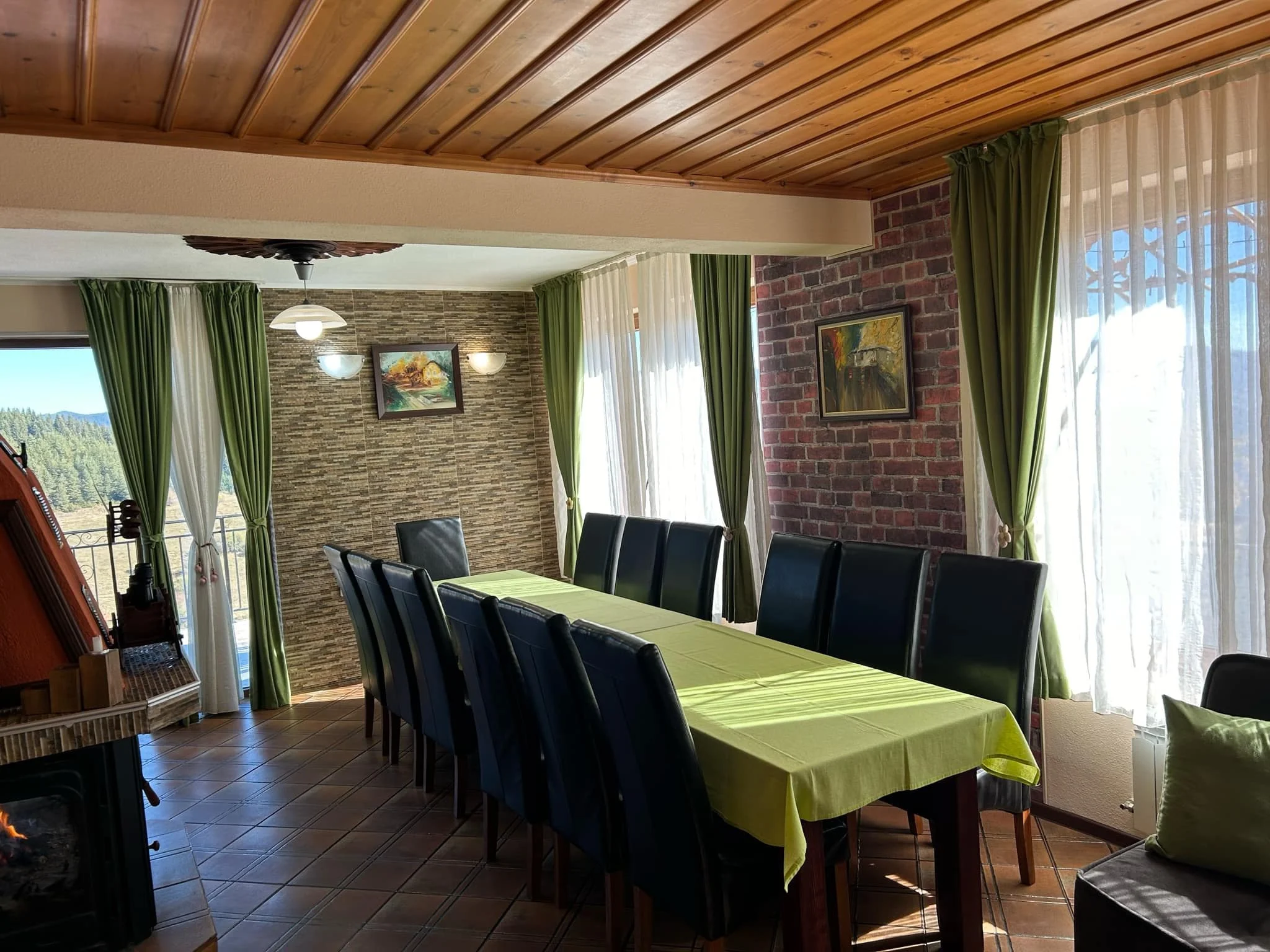 Dining room with black chairs around a table covered with a lime green tablecloth, large windows with green curtains, a brick wall, paintings, and a ceiling with wood paneling.