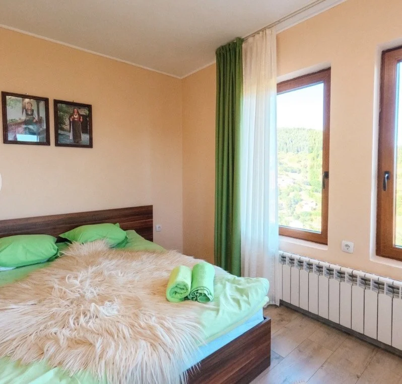 Bedroom with a bed, green pillows, a beige fur blanket, and rolled green towels. Two picture frames on the wall and two windows with green and white curtains showing a landscape outside.