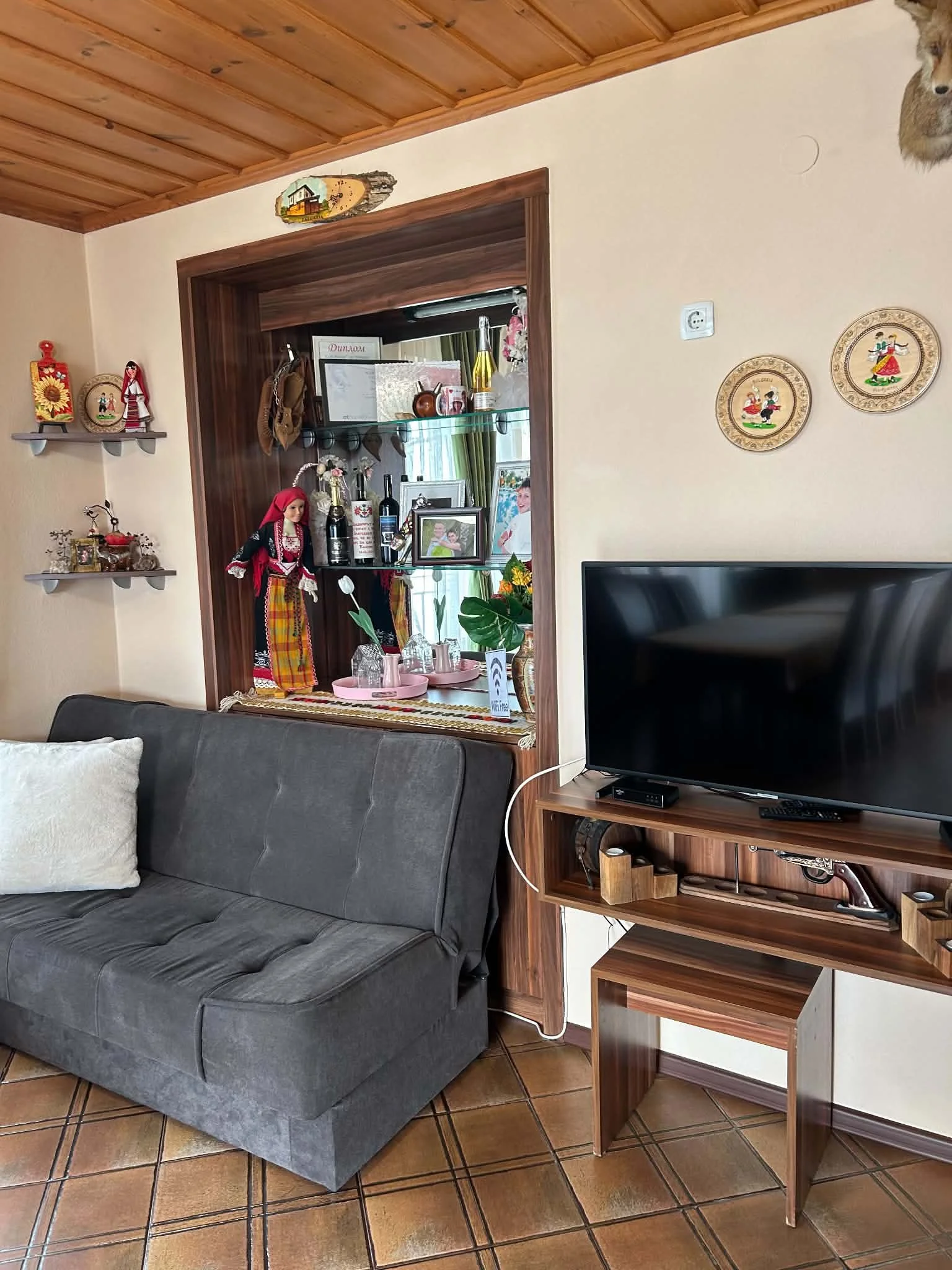 Living room corner with a dark grey fabric sofa, a wooden TV stand with a large flat-screen TV, and a wooden shelf with decorative items. The wall has three round decorative plates and part of a mounted animal head. There is a window with green curta