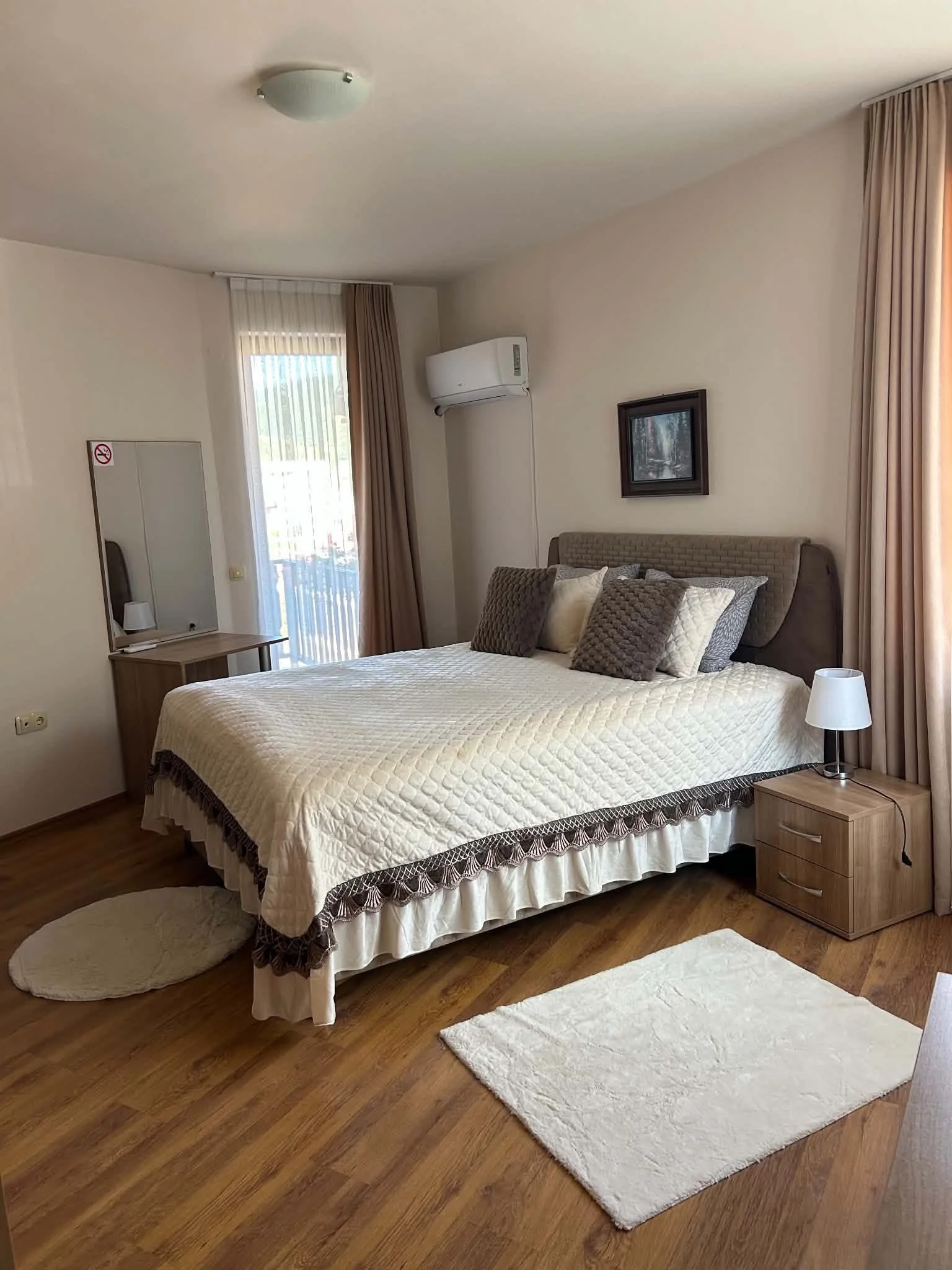 A hotel room with a bed, nightstand, lamp, wall art, air conditioning unit, window with curtains, and a rug on hardwood floor.