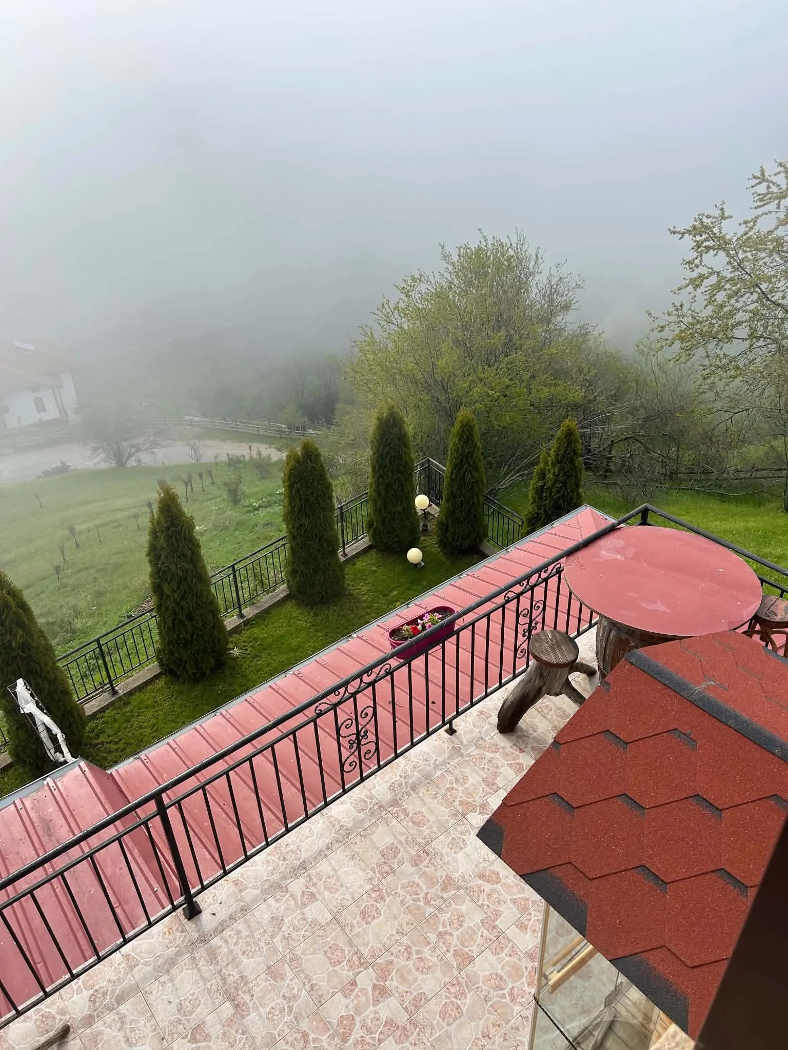 View from a balcony showing a wrought-iron railing, a round red table, a small wooden bench, and a terracotta roof. In the background, there are green trees, a grassy yard, and foggy weather obscuring the distant landscape.