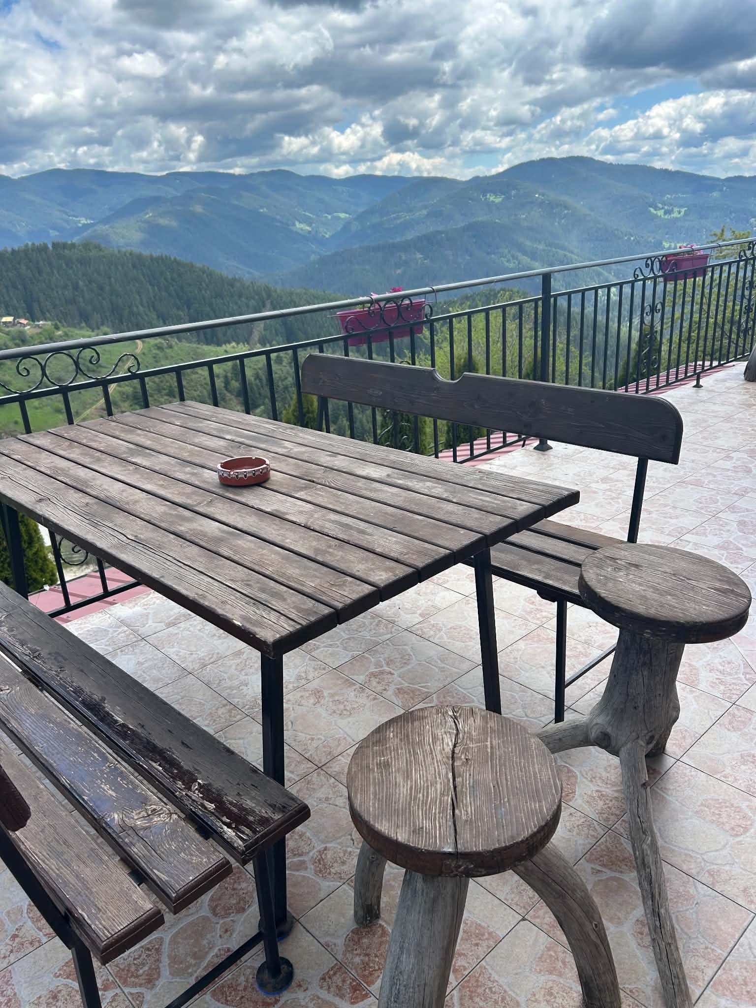 A wooden table with a small dish, surrounded by wooden and metal chairs on a tiled terrace overlooking a lush, green mountainous landscape under a partly cloudy sky.