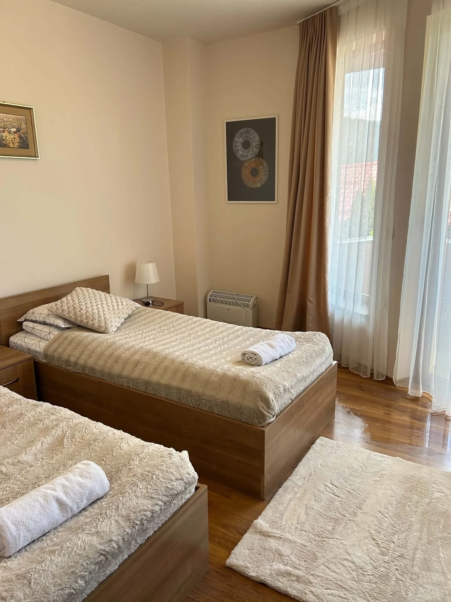 A bedroom with two single beds, each covered with a beige textured blanket, and a pillow. The beds are made of wood. There is a nightstand with a small white lamp and a rolled towel. The room has wooden flooring, cream-colored walls, and sheer curtai