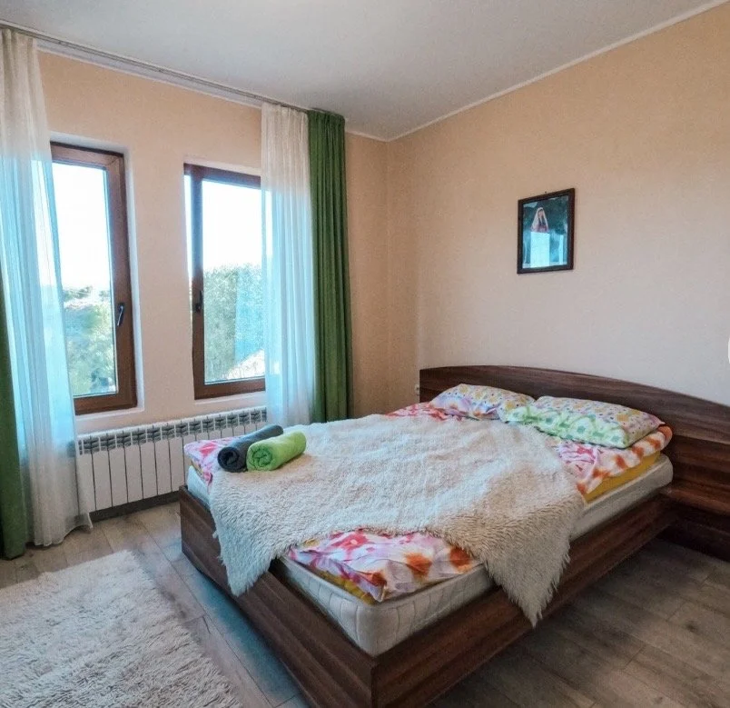 A bedroom with two windows, a wooden bed with colorful sheets and pillows, a blanket, and two rolled towels on the bed. There are white and green curtains and a framed picture on the wall.