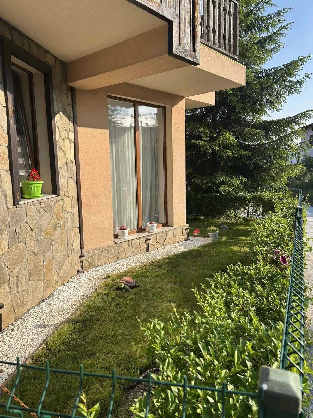 The image shows a house with a stone and beige exterior, large windows with curtains, and a small garden with green bushes, colorful flowers, and a pathway lined with white pebbles. There are also potted plants on the windowsill and small garden orna